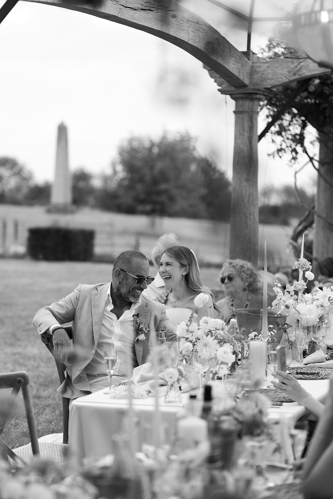 Black and white photo of a couple sharing a joyful moment at an elegantly decorated outdoor wedding reception table.