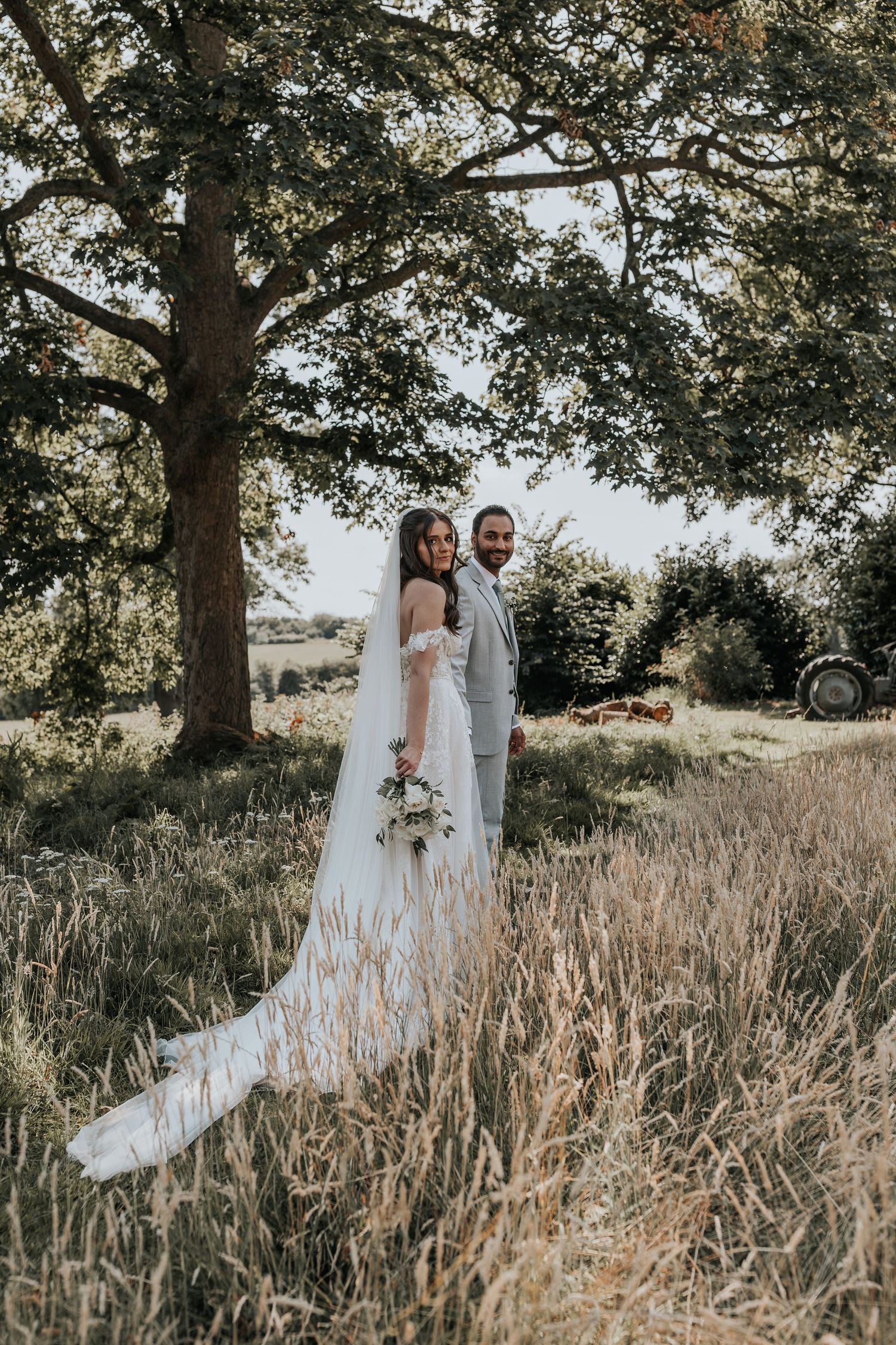 Bride and groom posing in a golden meadow under a large tree on their wedding day.