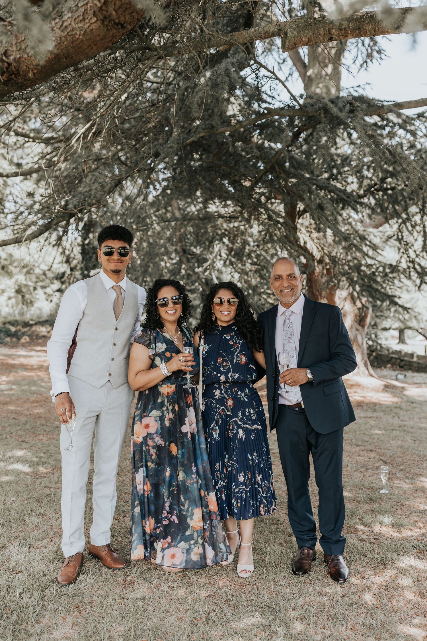 Four elegantly dressed guests posing together outdoors under a large tree at a wedding celebration.