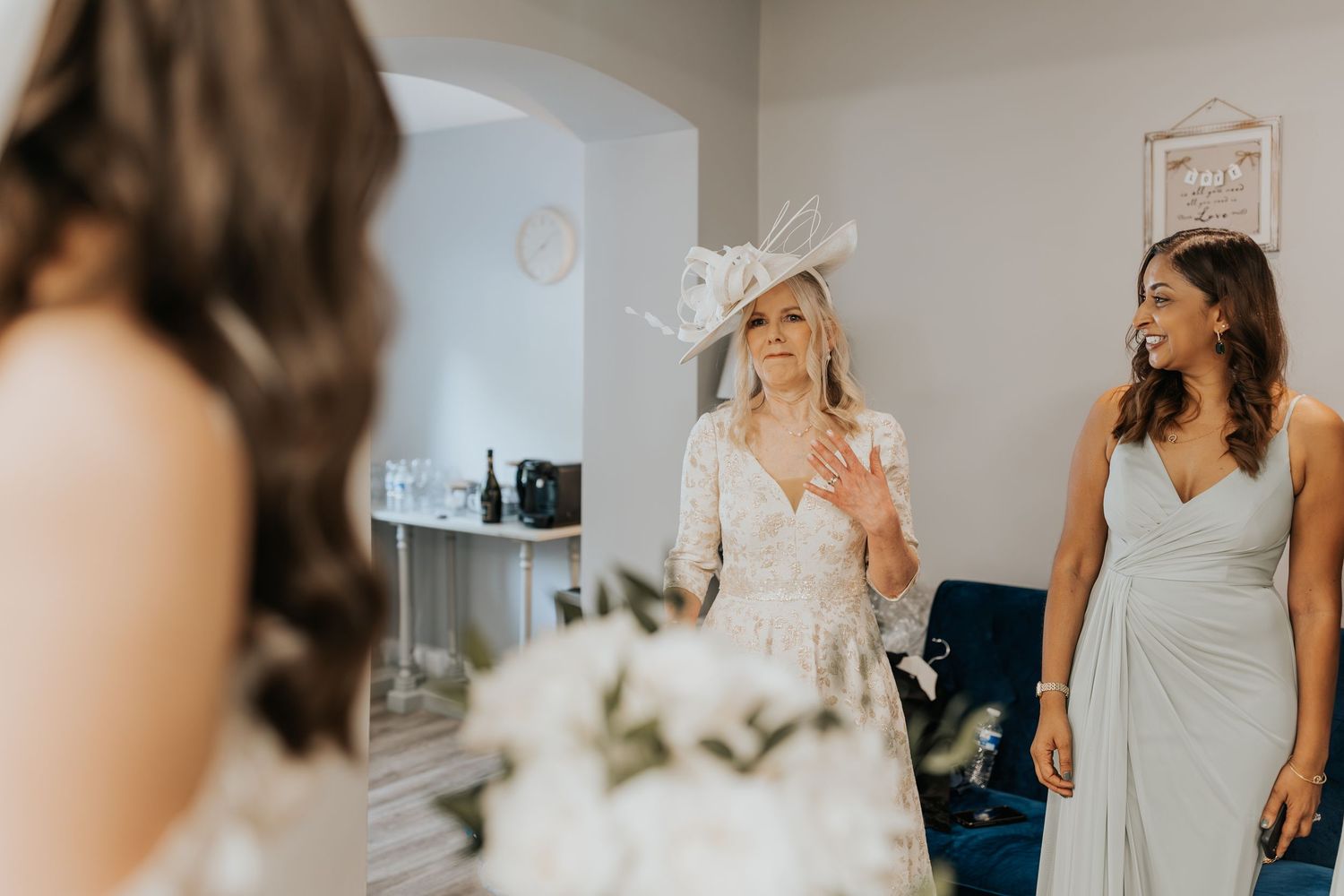 Bride in lace gown holds white bouquet as guests in elegant attire react with joy during wedding preparations.