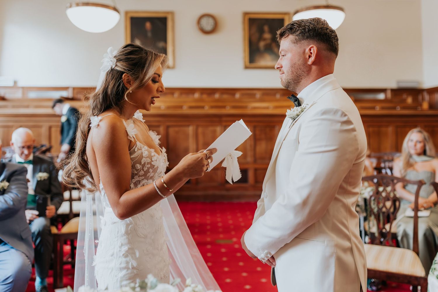Bride reading wedding vows to groom in white suit during indoor ceremony.