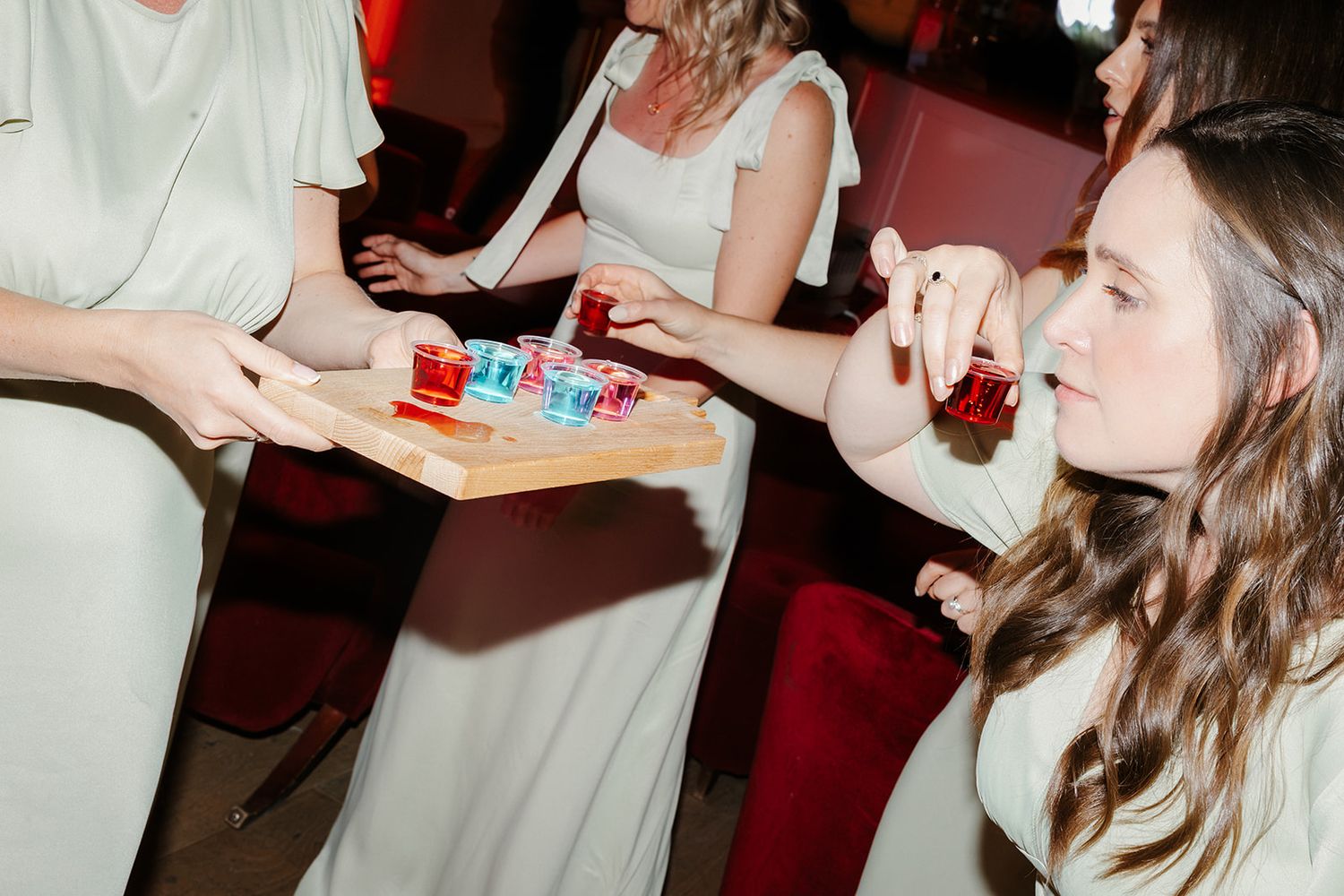 Women in white dresses taking colorful shots from a wooden tray at a wedding reception party.