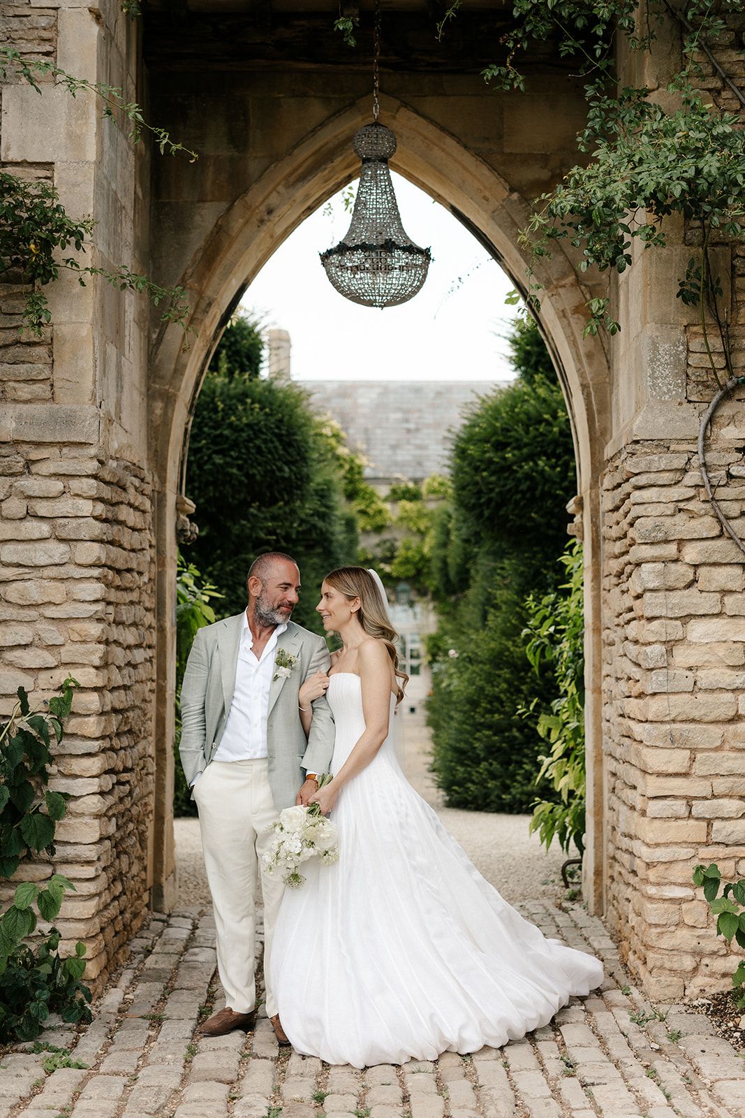 Bride and groom stand under a stone arch with chandelier, surrounded by lush gardens at an elegant wedding venue.