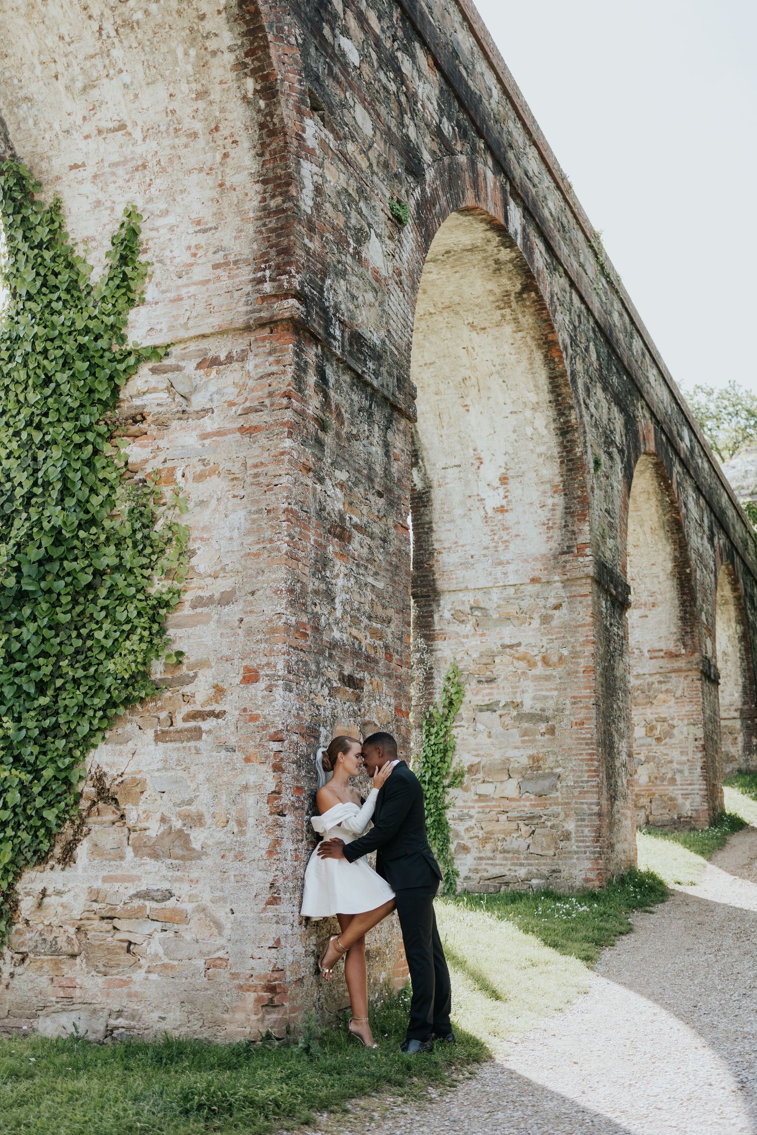 Couple in wedding attire embracing against a historic brick arched aqueduct wall with ivy, outdoor romantic photo.