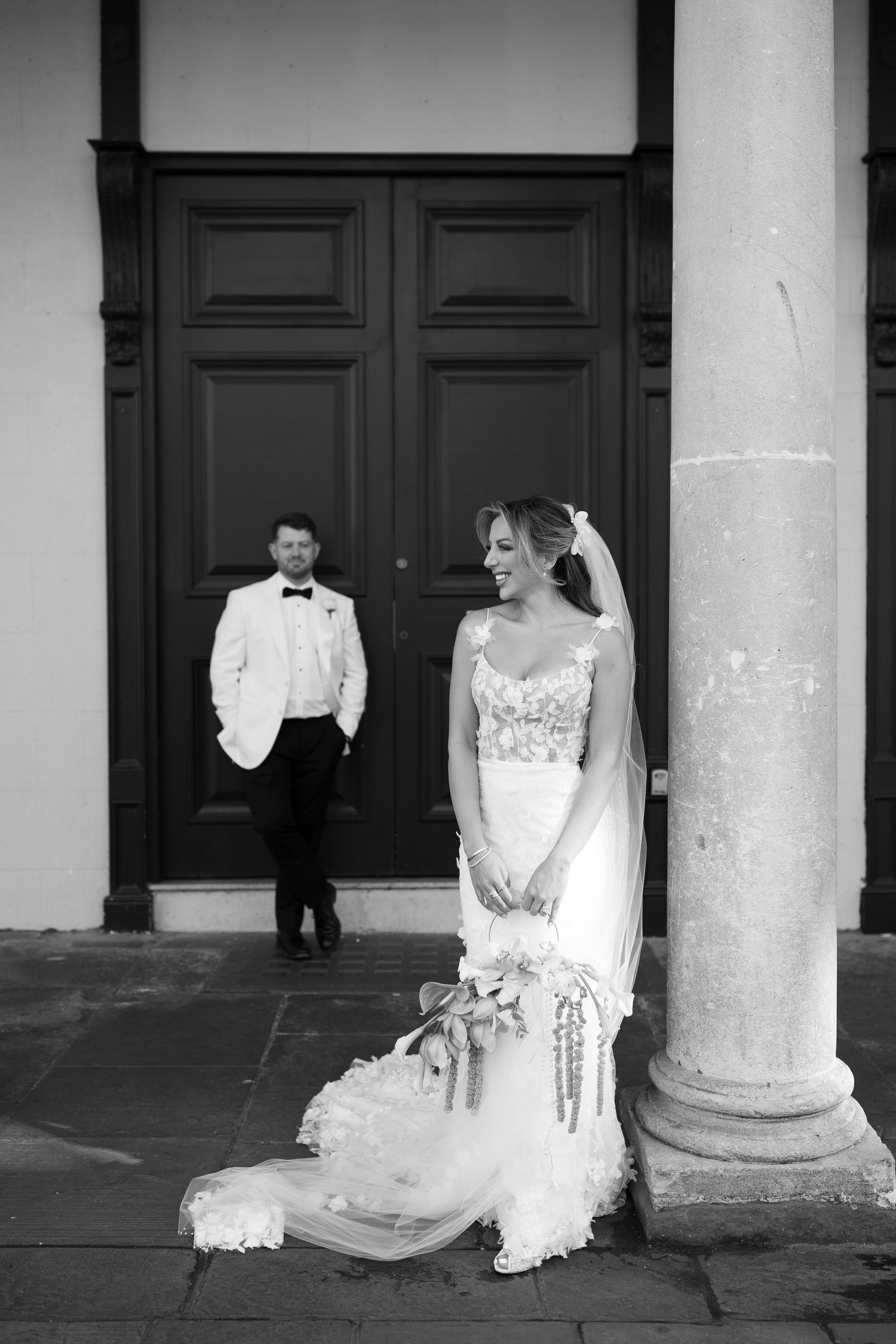 Black and white wedding photo of bride in lace gown leaning on column, groom in white tuxedo by grand doors.