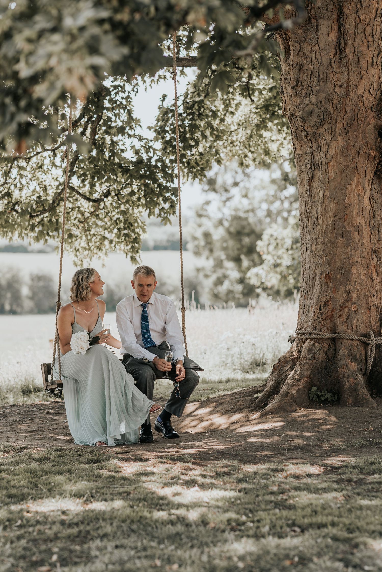 A bride and groom sit together on a wooden swing hanging from a large tree outdoors on their wedding day.