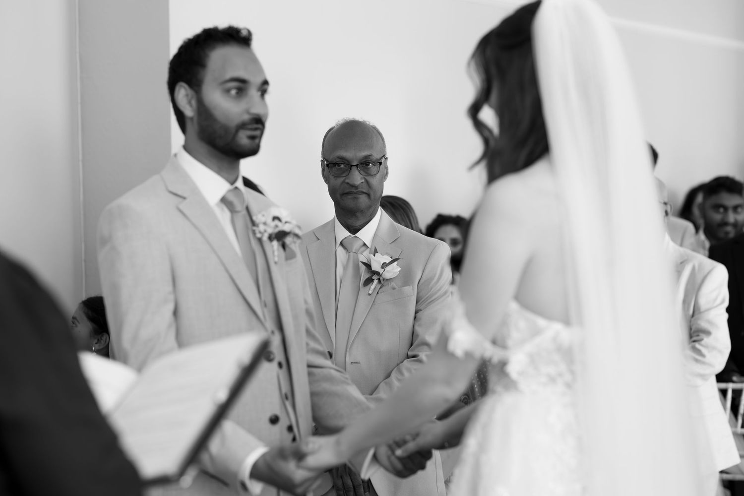 Black and white wedding ceremony photo of groom holding bride's hands at the altar with guests seated behind.