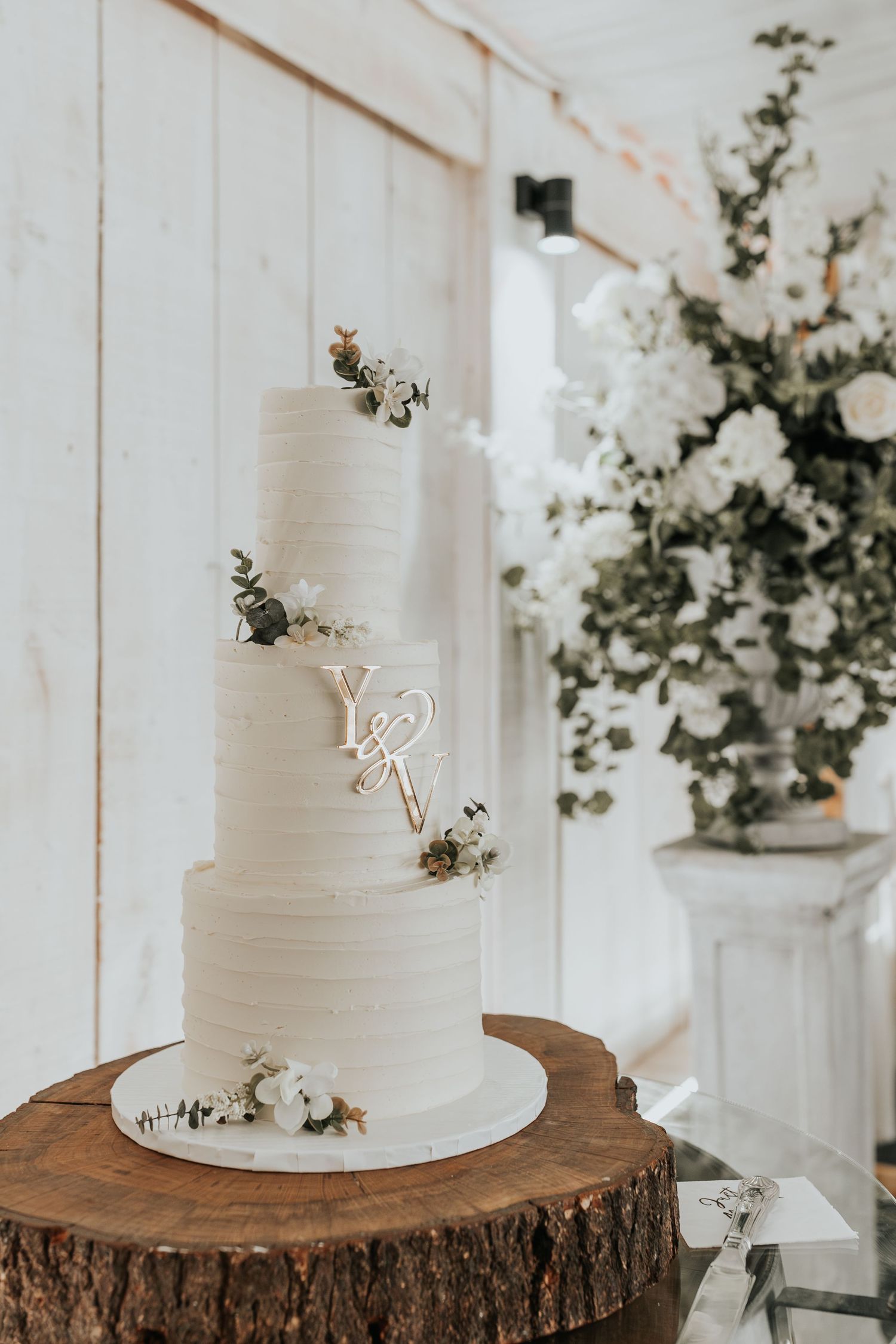 Three-tier white textured wedding cake with gold monogram initials and floral accents displayed on a rustic wood slice.