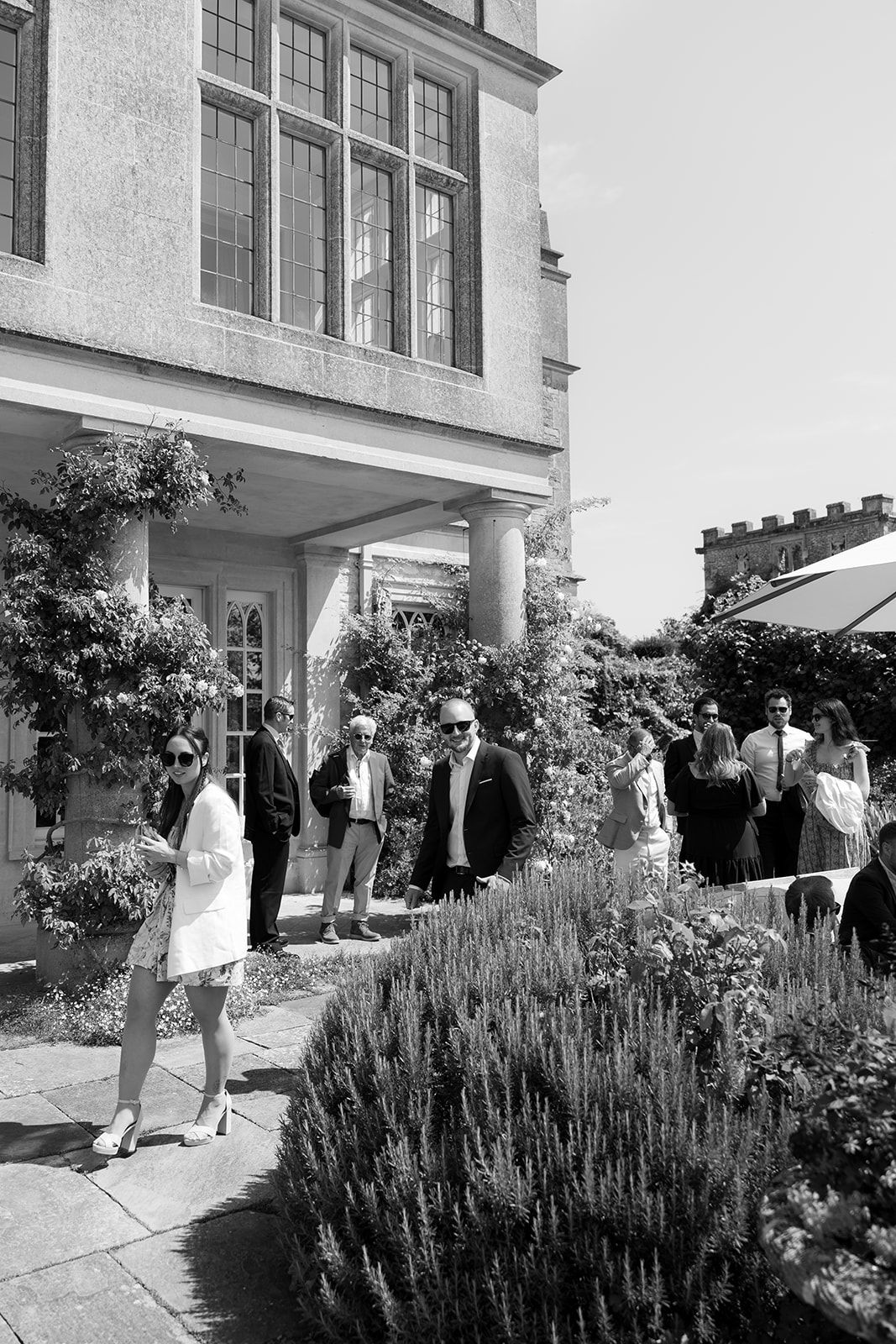 Black and white photo of elegantly dressed guests socializing outside a grand country house on a sunny day.
