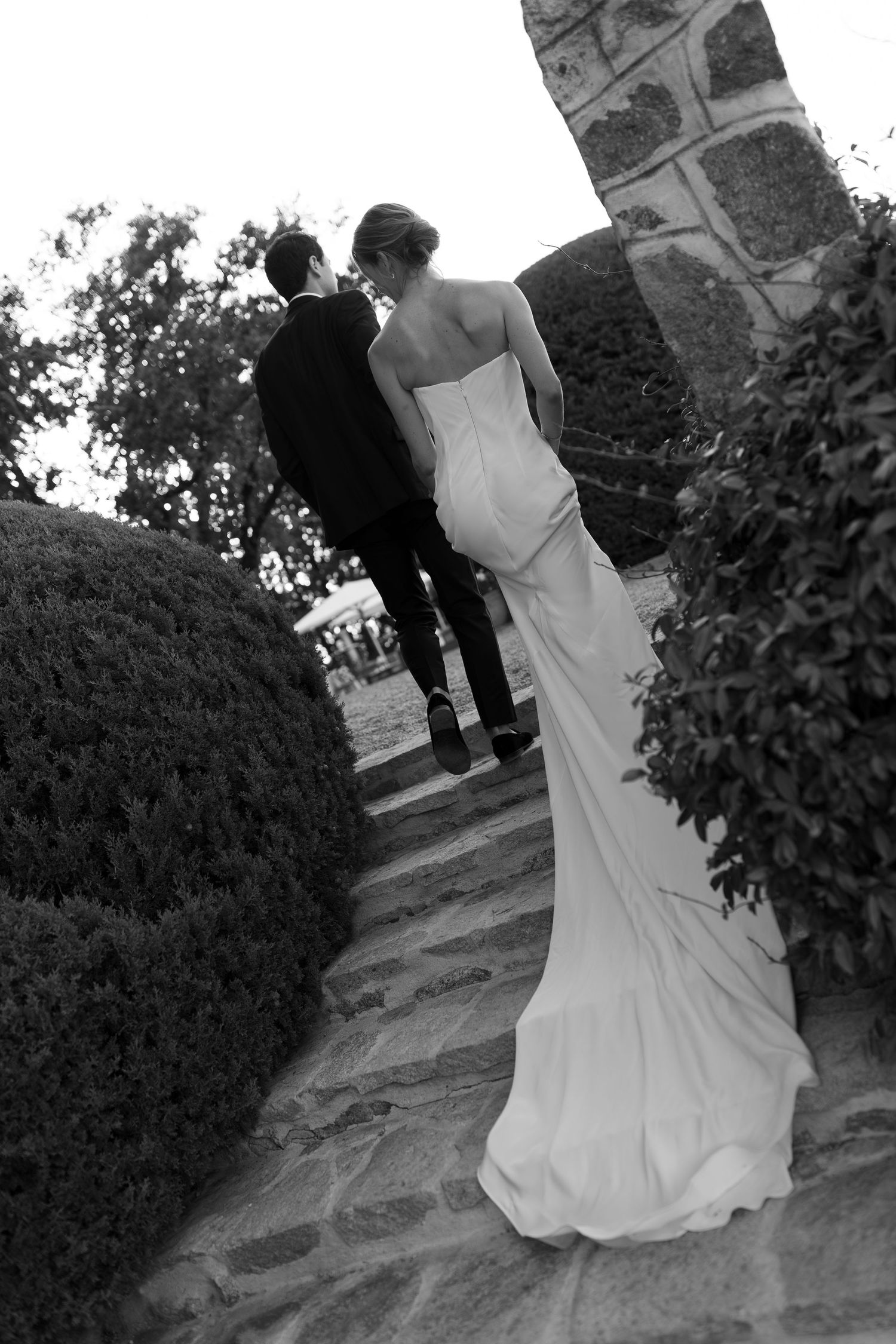 A black and white photograph of a bride and groom walking together on a stone path surrounded by greenery.
