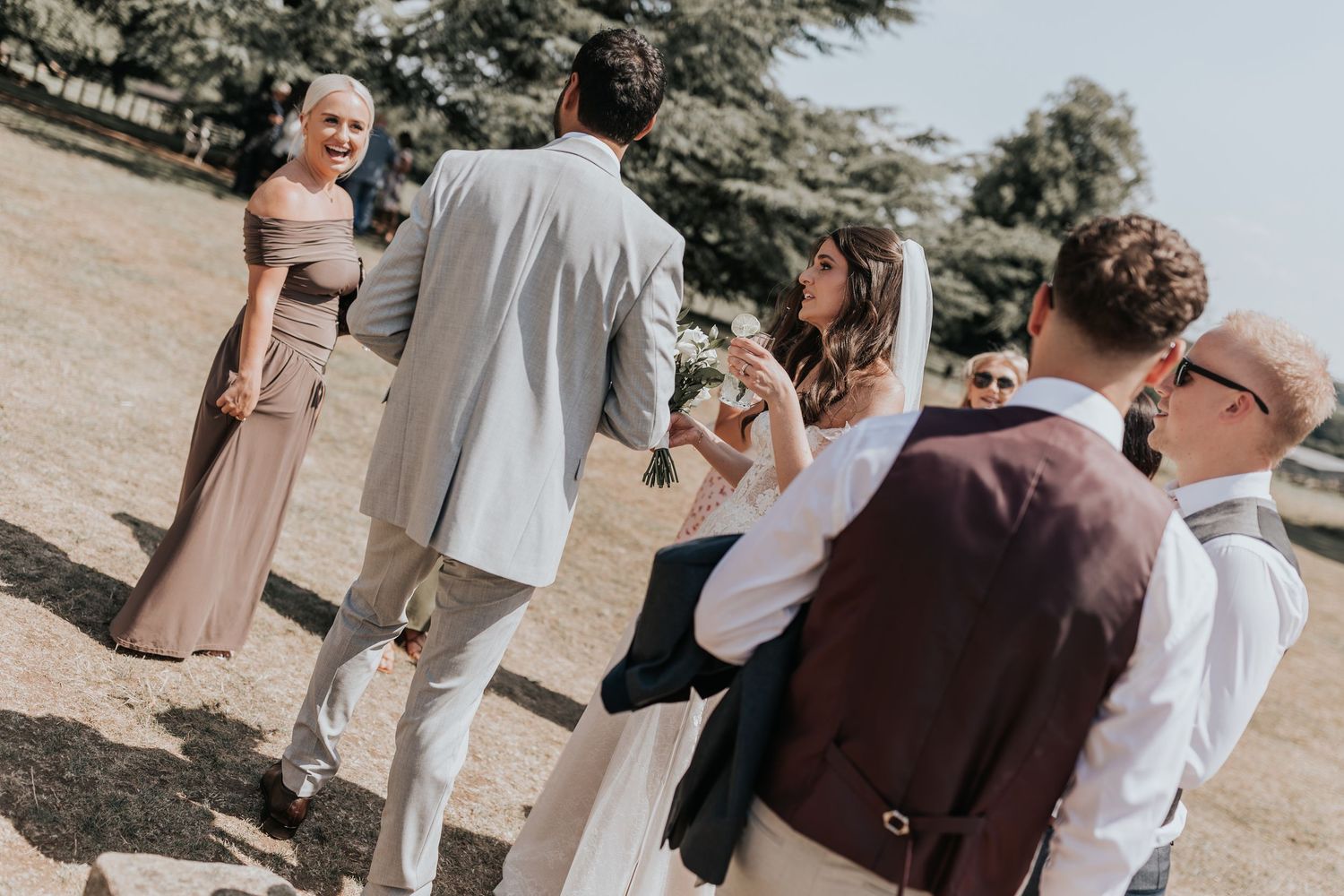Wedding party guests in formal attire socialising outdoors on a sunny day, with bride holding bouquet.