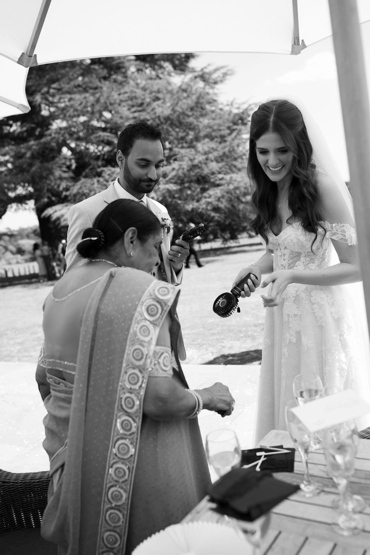 Black and white wedding photo of bride in lace gown with groom and elder woman in sari during outdoor ceremony.