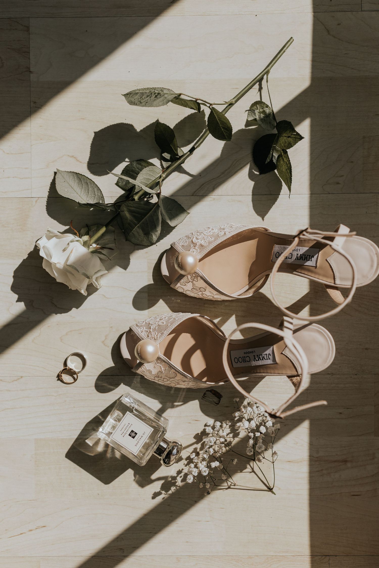 Flat lay of bridal accessories including Jimmy Choo heels, pearl earrings, gold rings, Jo Malone perfume, and a white rose.
