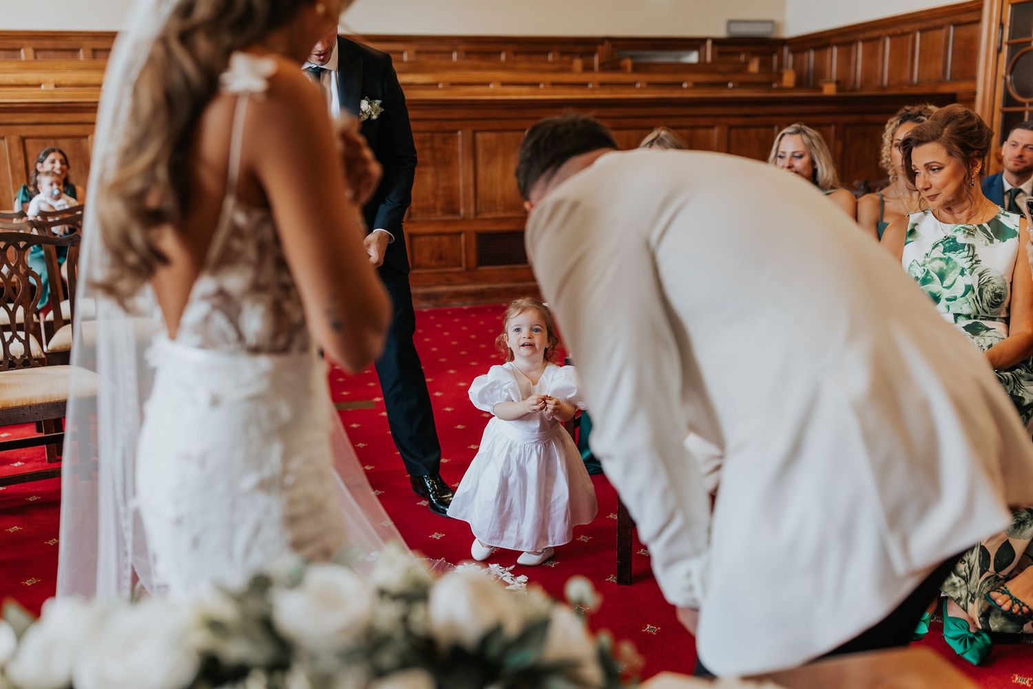 A flower girl in white dress walks toward groom bending down to greet her during elegant church wedding ceremony.