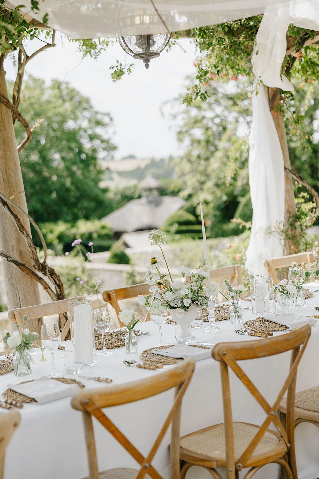 Elegant outdoor wedding reception table with white linens, floral centerpieces, and wooden cross-back chairs surrounded by lush greenery.