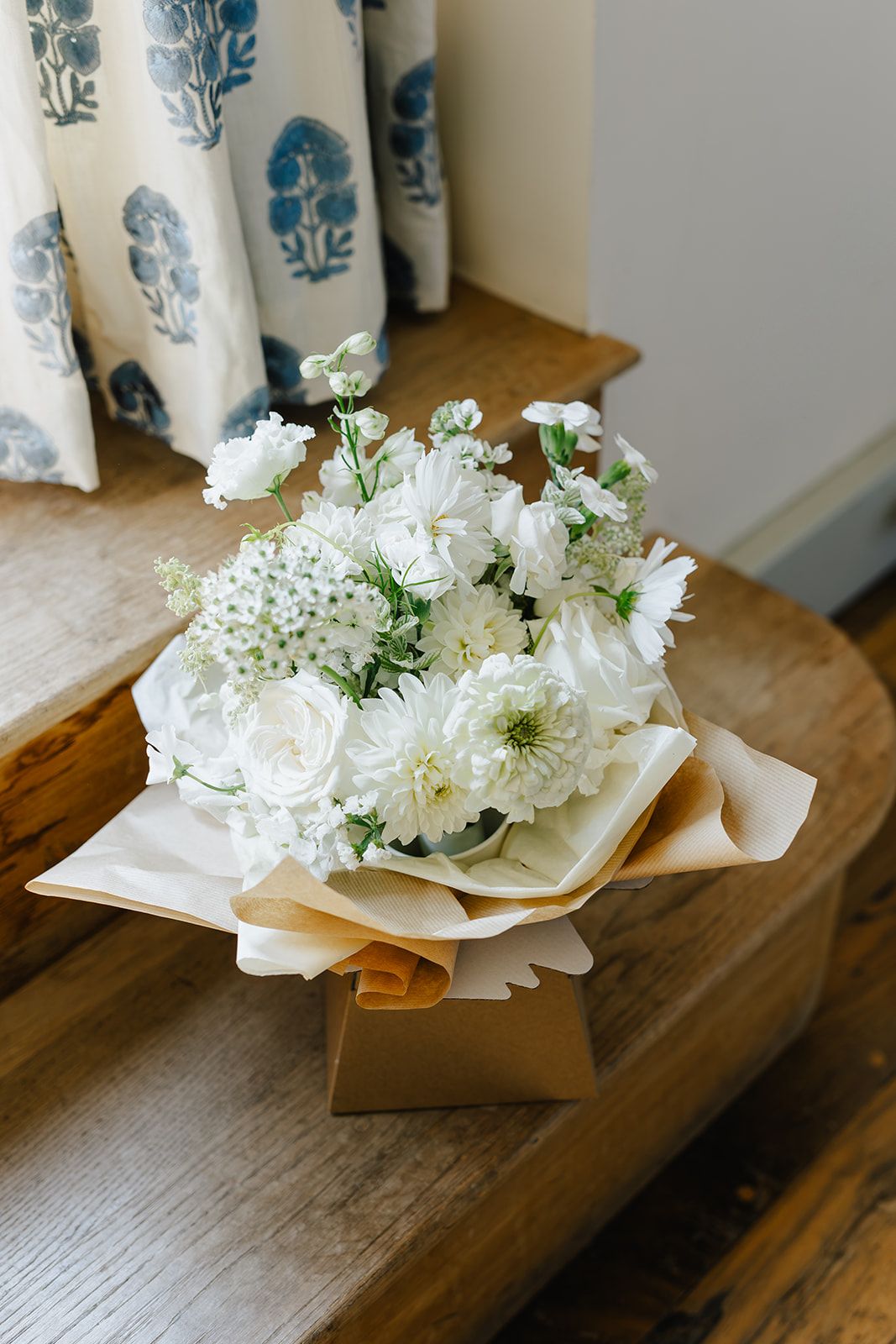 White floral bouquet with roses and dahlias wrapped in kraft paper, displayed on a wooden bench near blue floral curtains.
