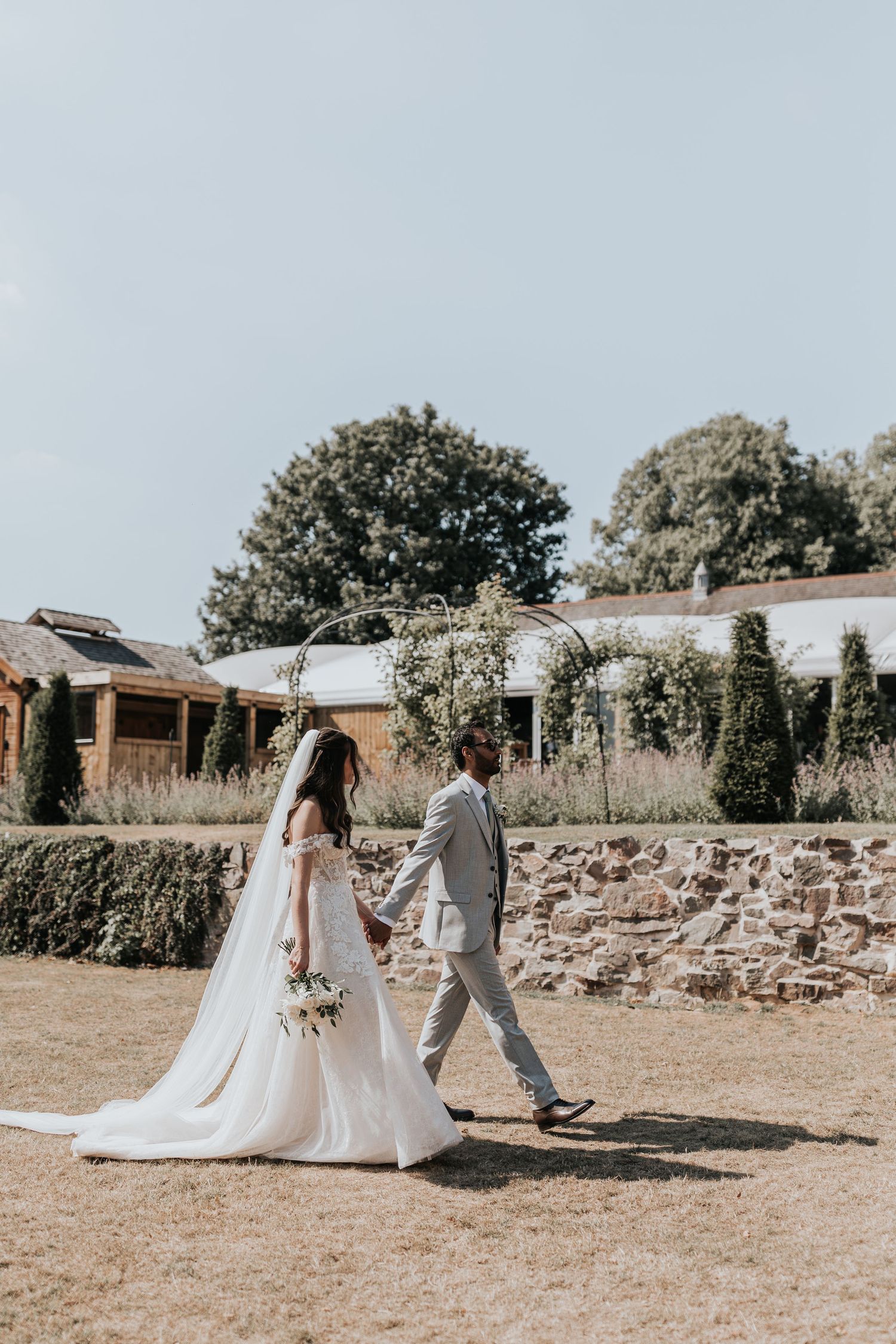 A bride and groom walk hand in hand through a sunny garden venue, she in a flowing white gown, he in a light gray suit.