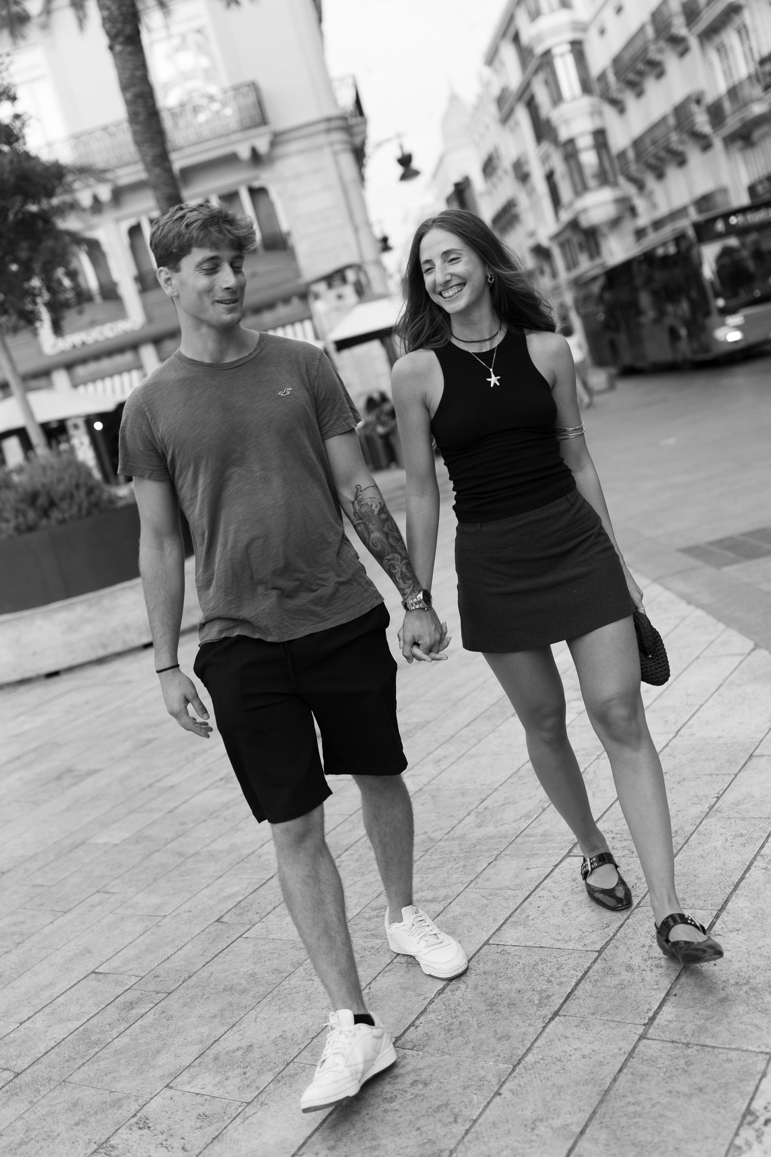 A young couple walks hand-in-hand down a city street, dressed casually and smiling.