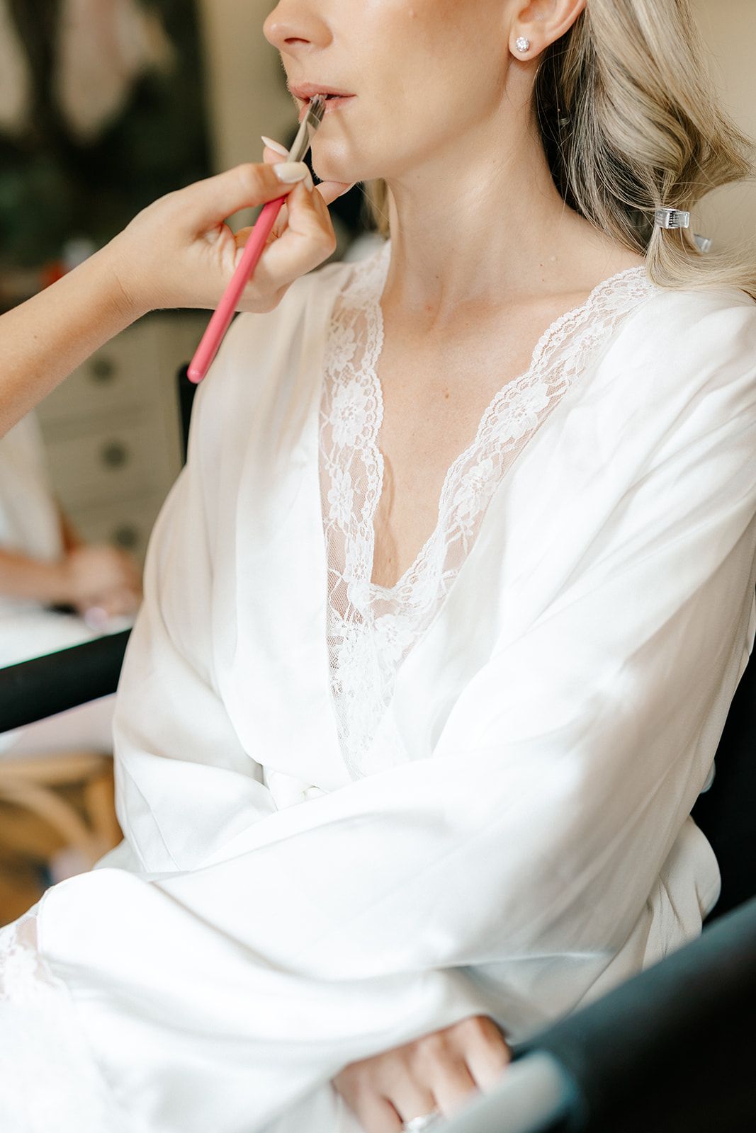 Bride in white satin robe getting lip makeup applied by makeup artist during wedding morning preparation.