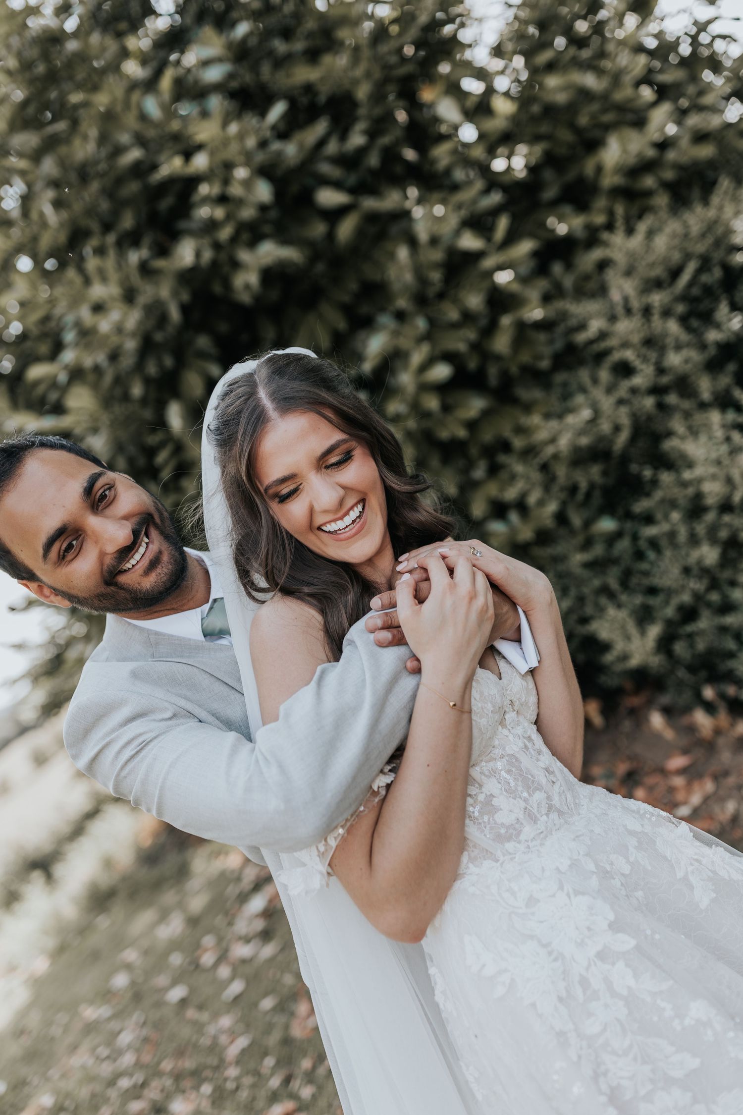 A joyful bride and groom embrace outdoors, laughing together in front of lush green hedges on their wedding day.