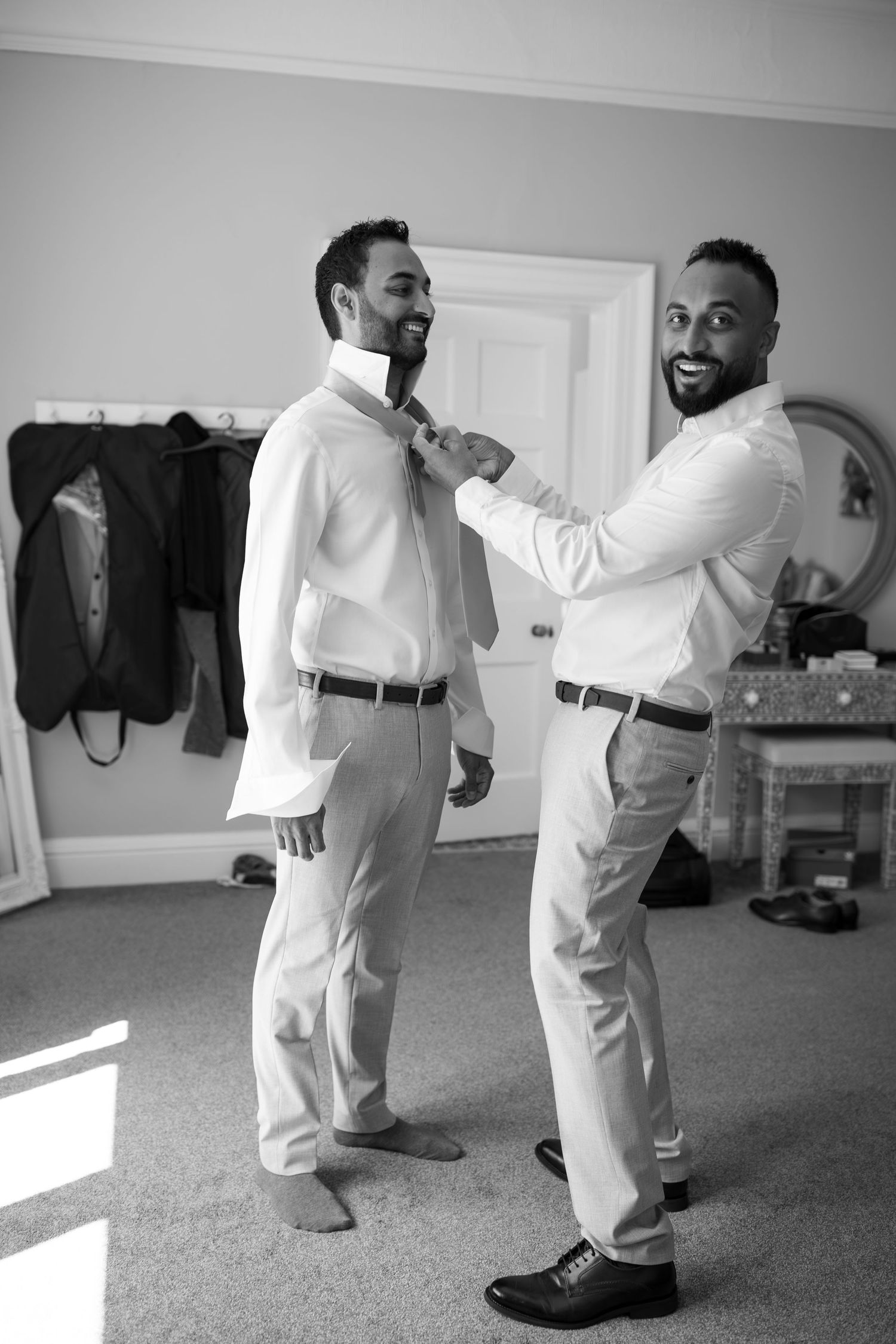 Black and white photo of two men in dress shirts, one helping the other adjust his tie while smiling in a bright room.