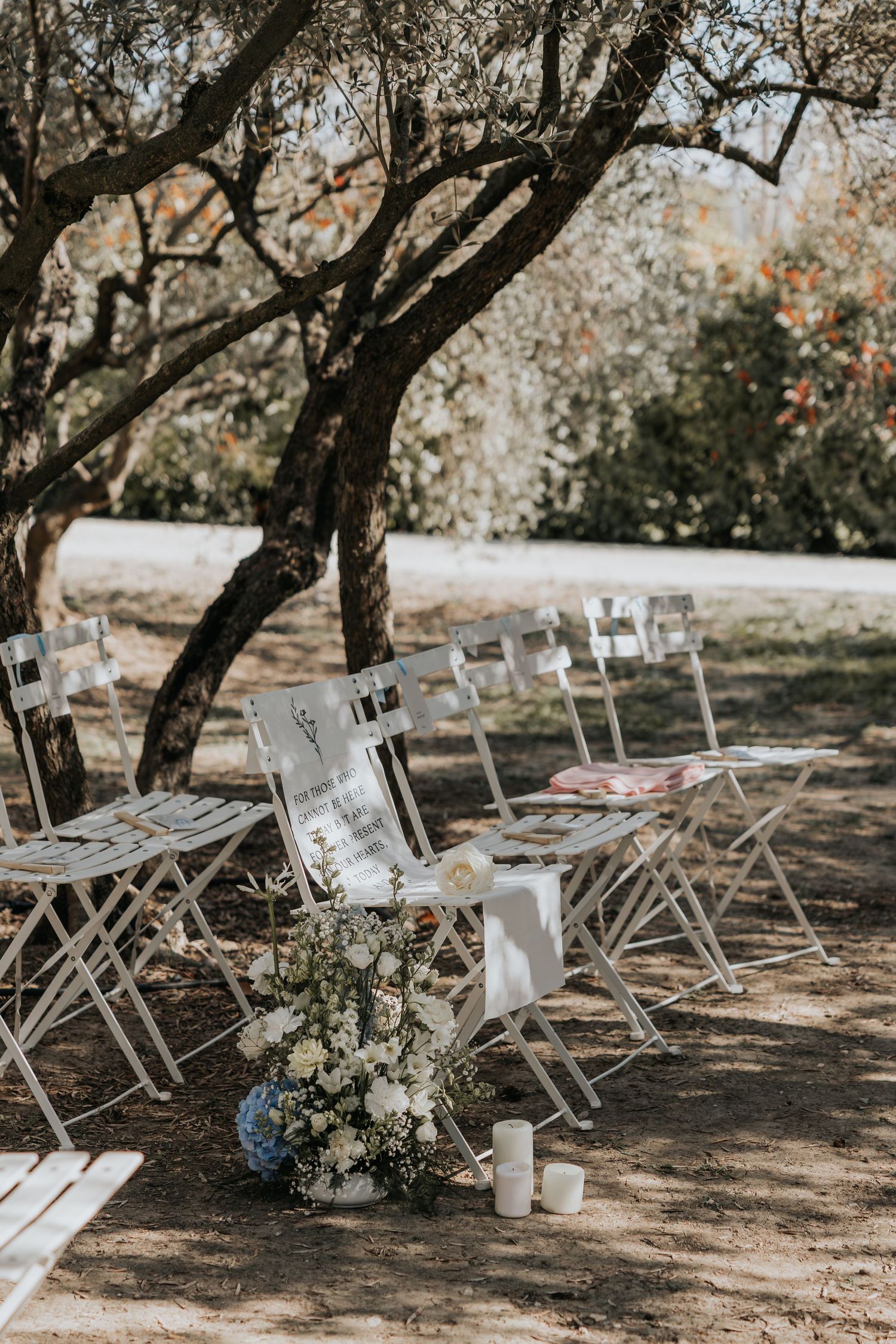 A rustic outdoor wedding ceremony setup with wooden chairs and a floral arrangement under a canopy of trees.