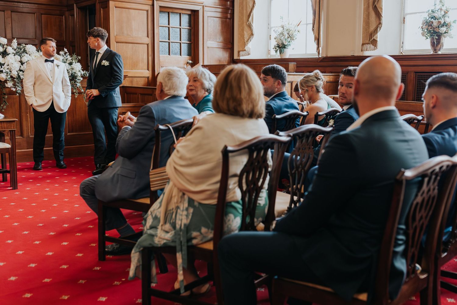 Guests seated in elegant wood-paneled wedding ceremony room with red carpet, watching groom at altar.
