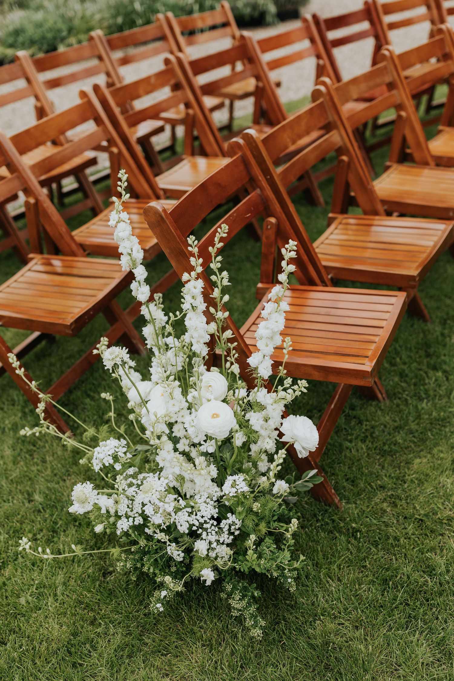 White floral arrangement with roses and delphinium beside wooden folding chairs on green grass at an outdoor wedding ceremony.