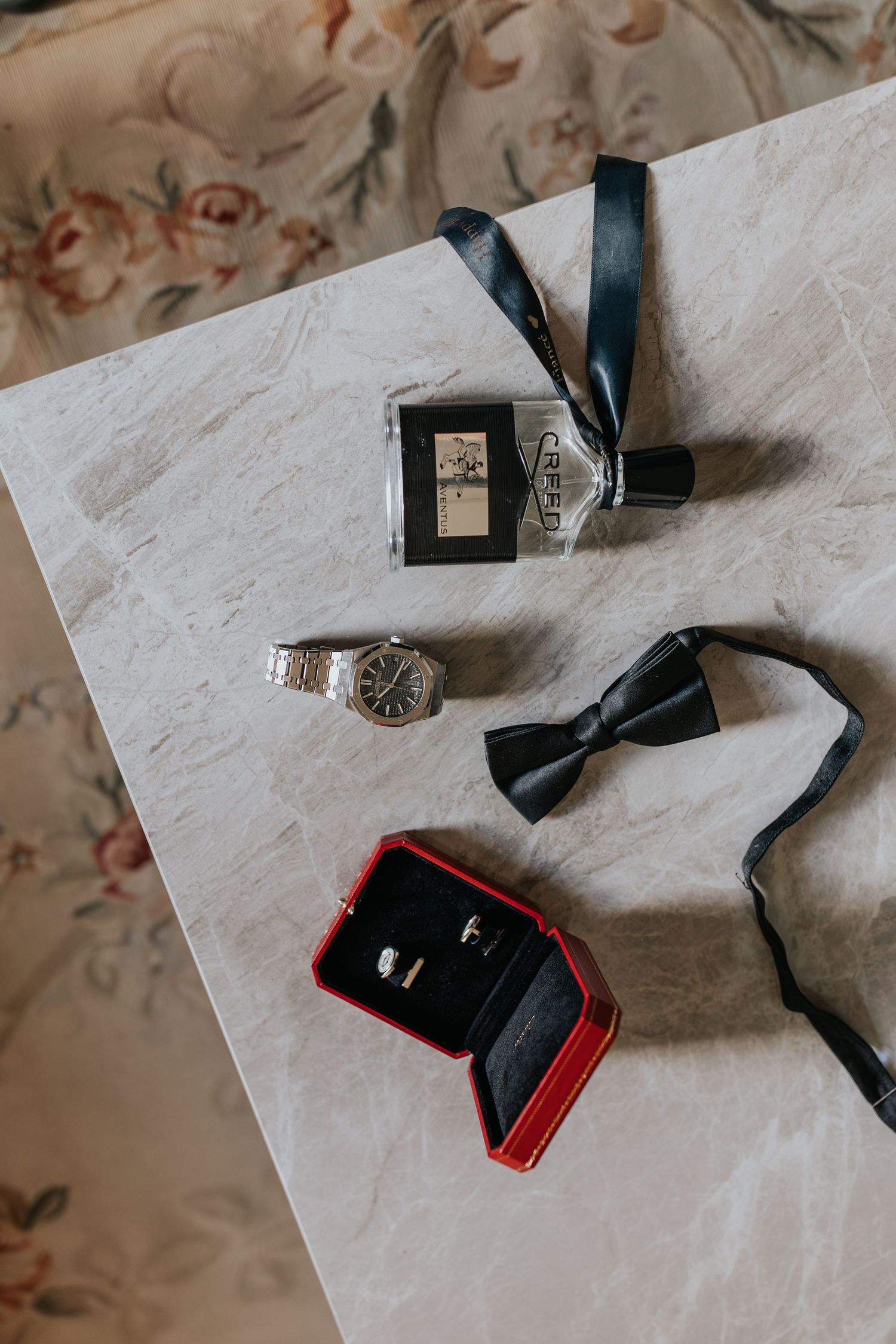 Groom wedding accessories flat lay featuring a silver watch, black bow tie, cologne, and wedding rings in a red velvet box.