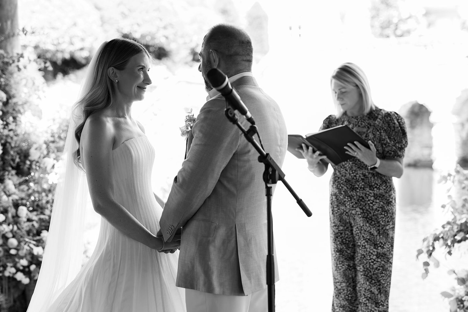 Black and white photo of a bride and groom exchanging vows at an outdoor wedding ceremony with an officiant.