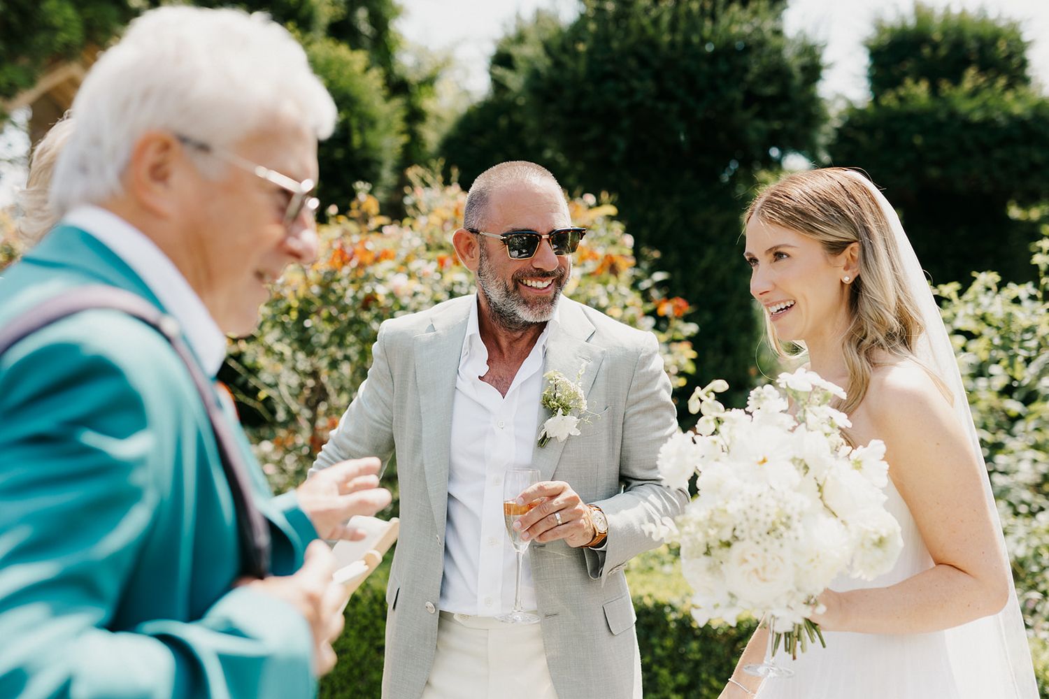 Bride holding white bouquet smiles with groom in sunglasses and elder guest at outdoor wedding ceremony.