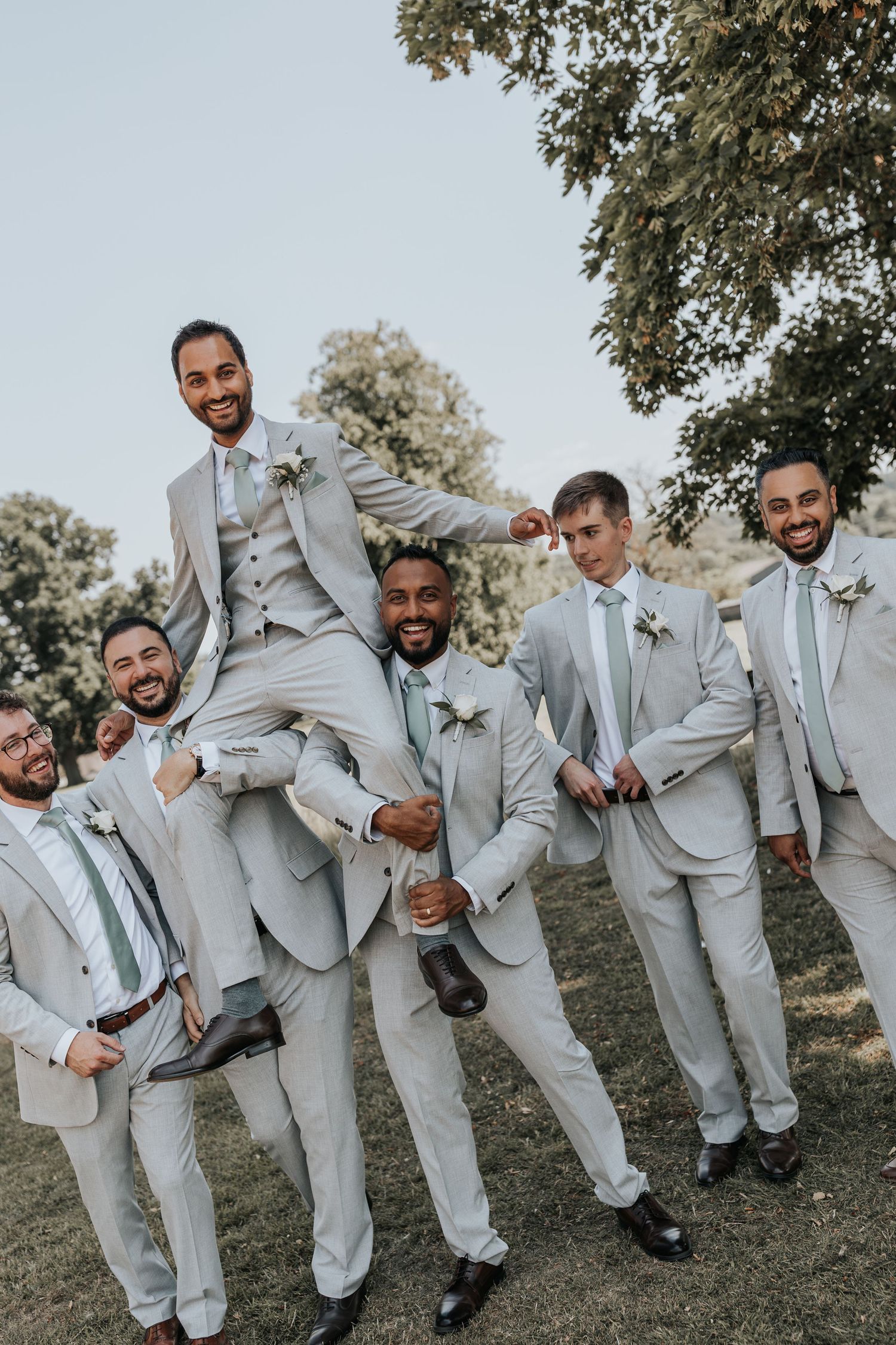 Groomsmen in light gray suits with sage green ties laugh and lift the groom outdoors at a wedding celebration.