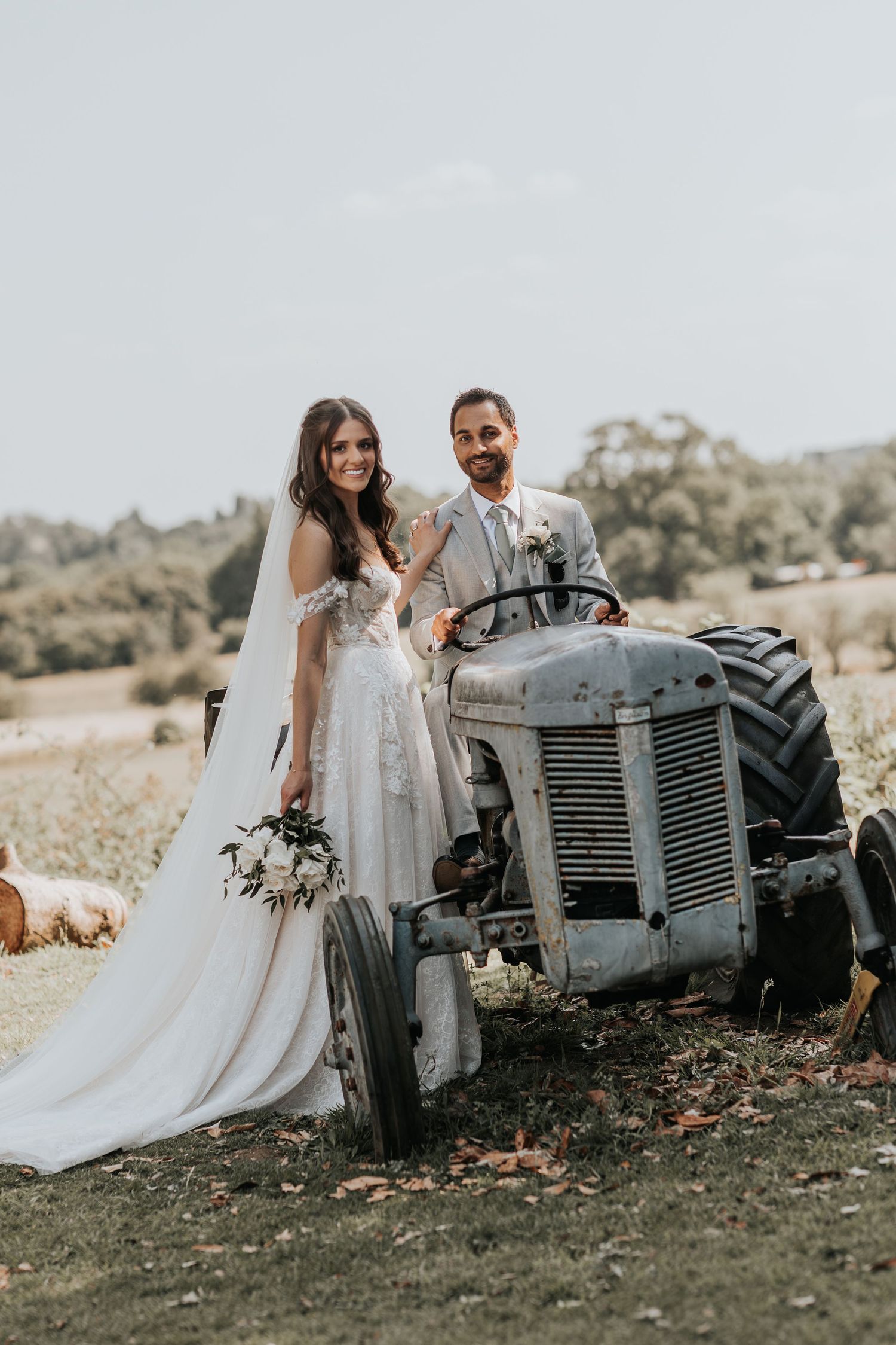 Bride in white gown holds bouquet beside groom on vintage tractor at rustic farm wedding outdoor setting.