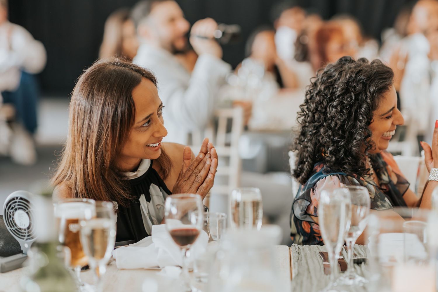 Two women smiling and clapping at an elegant event dinner, with wine glasses and table settings in the foreground.