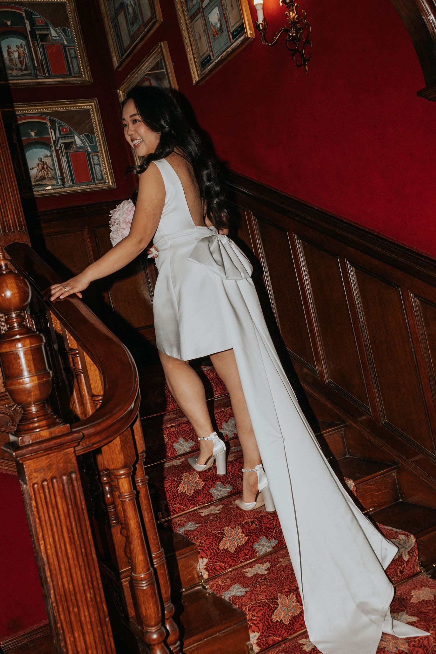 A woman in a white dress stands on a staircase with a red carpet, surrounded by wooden paneling and artwork.