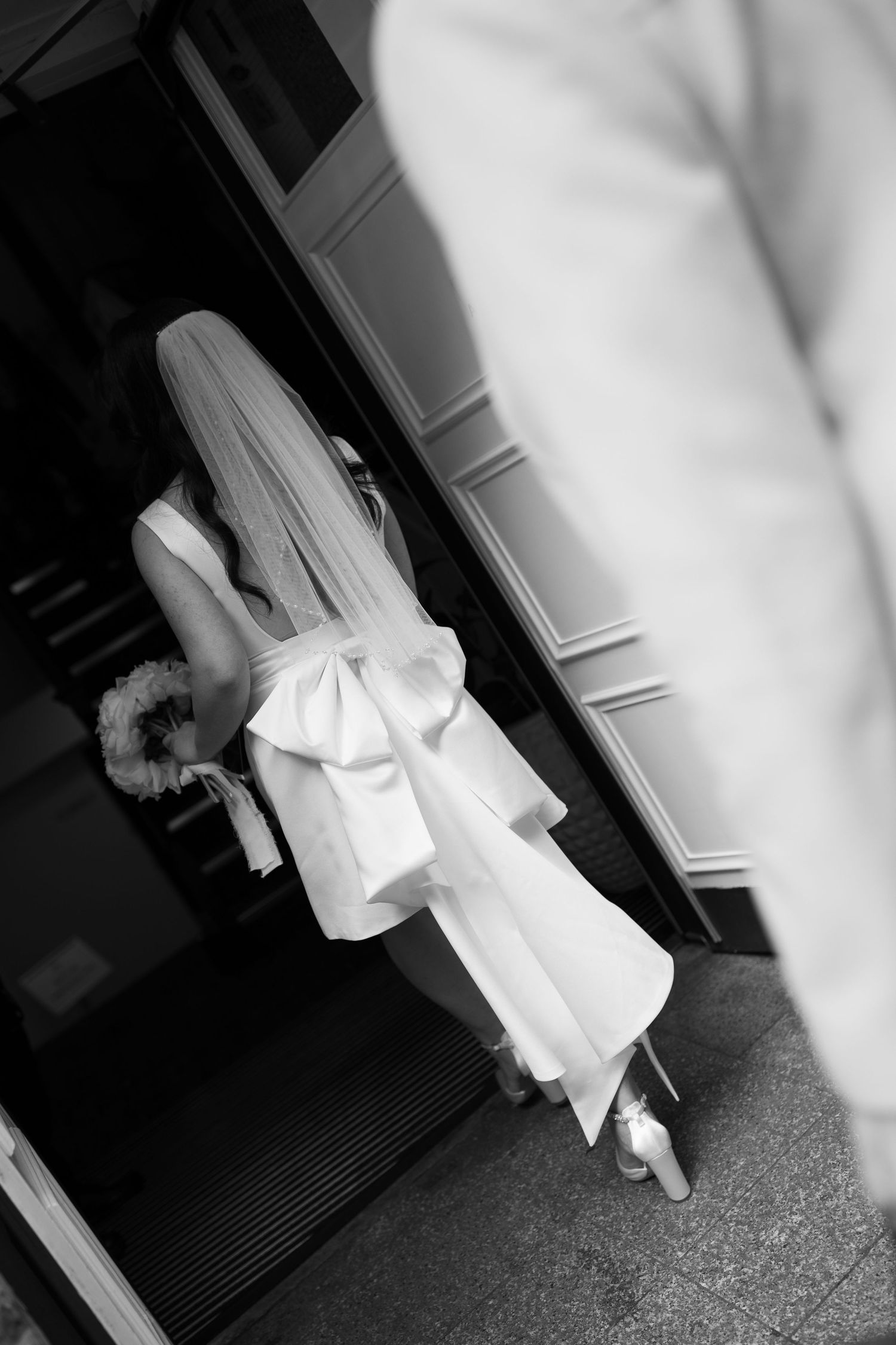 A black and white image of a woman wearing a long white dress with a bow, standing in a doorway.