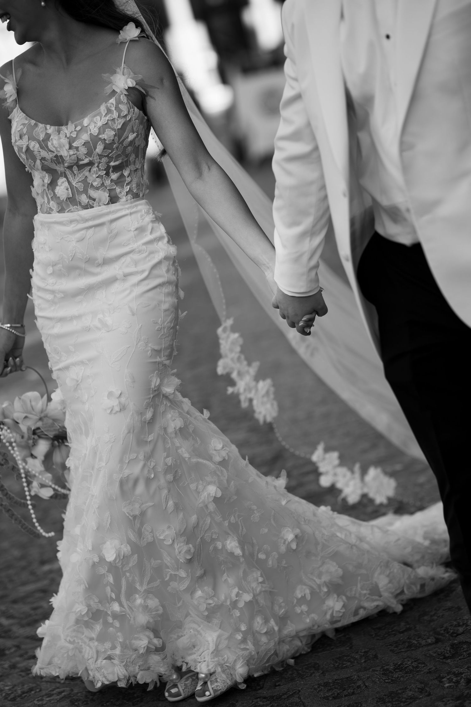 Black and white photo of bride in floral lace gown with train holding hands with groom in white tuxedo jacket.