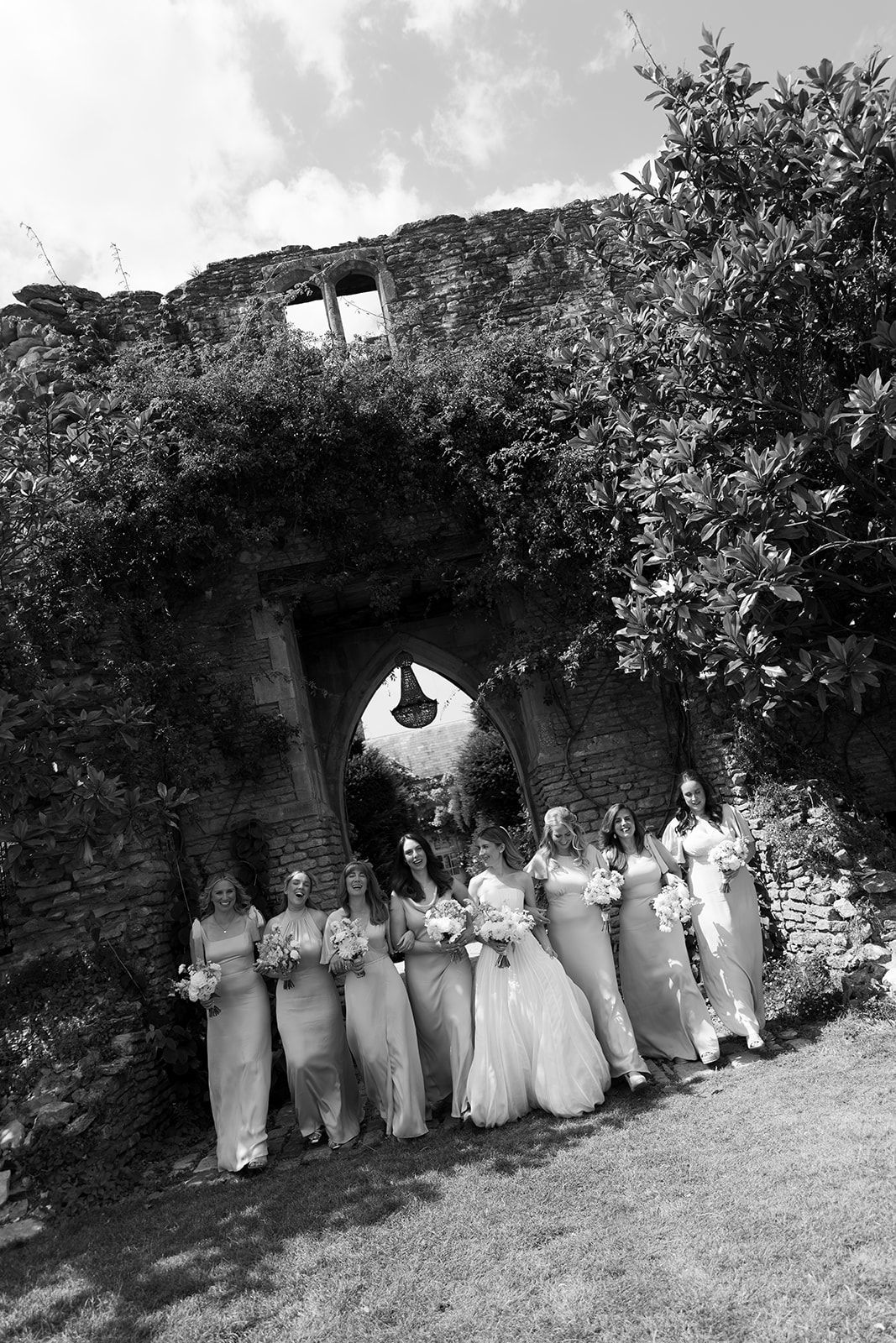 Black and white photo of a bride and bridesmaids posing in front of a rustic stone archway covered in ivy.