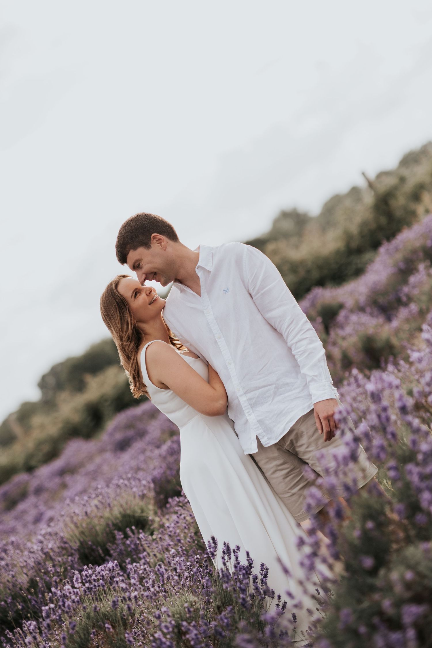 A couple embracing in a field of lavender flowers, with a scenic mountain backdrop.
