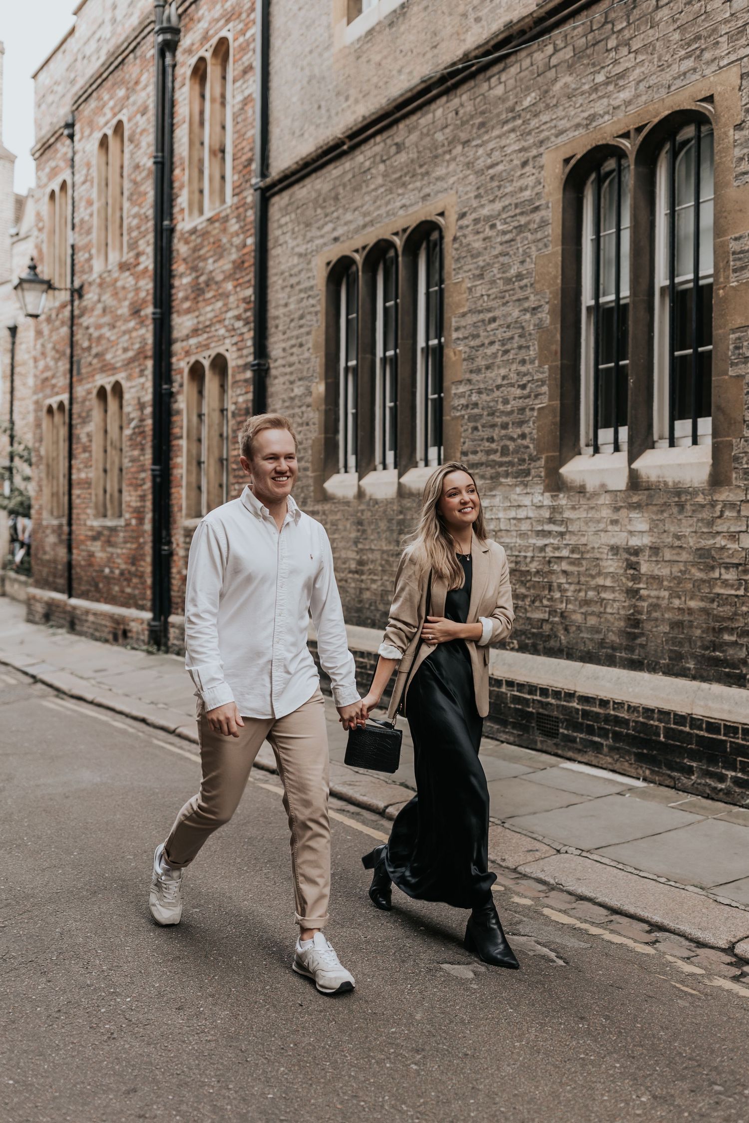 A couple walks together on a city street in front of a brick building with arched windows in Cambridge