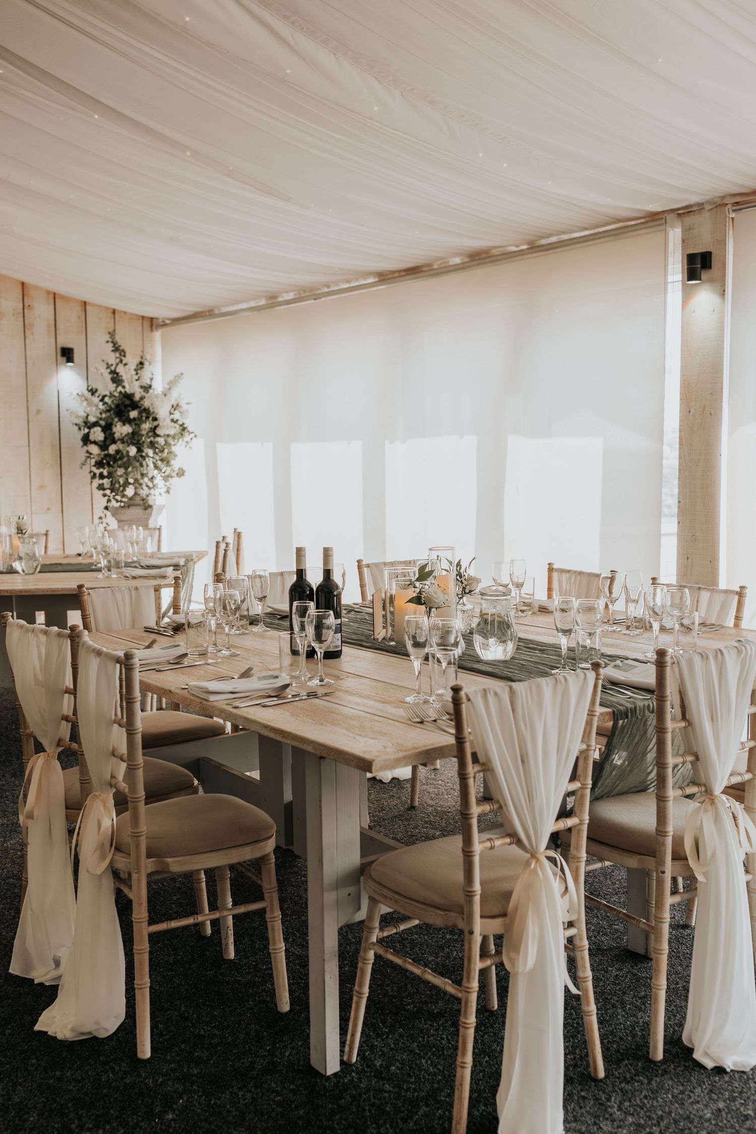 Elegant wedding reception marquee with draped ivory chairs, wooden tables, floral centerpieces, and soft white fabric ceiling decor.