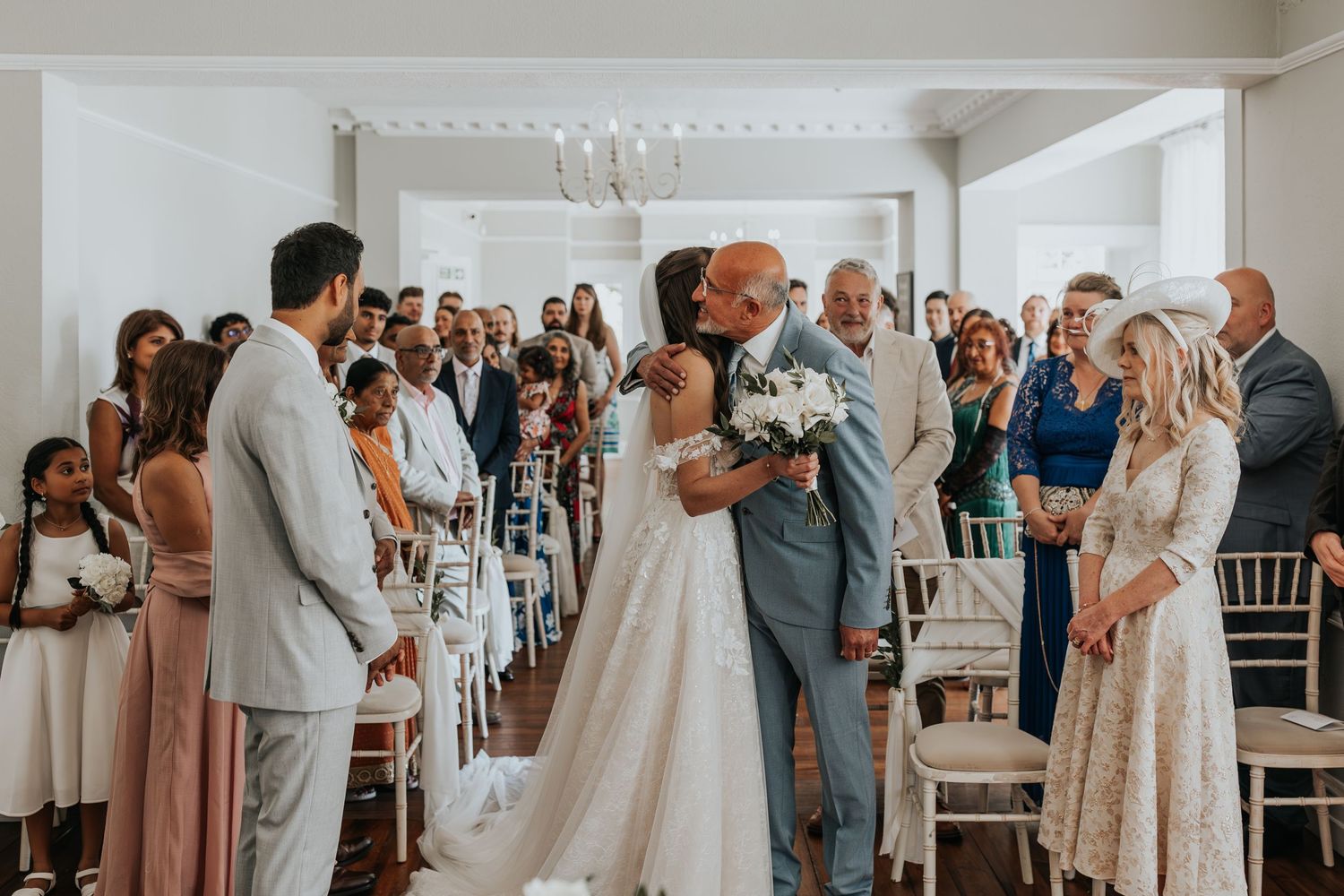Bride and groom share first kiss at wedding ceremony aisle surrounded by guests in elegant white venue.
