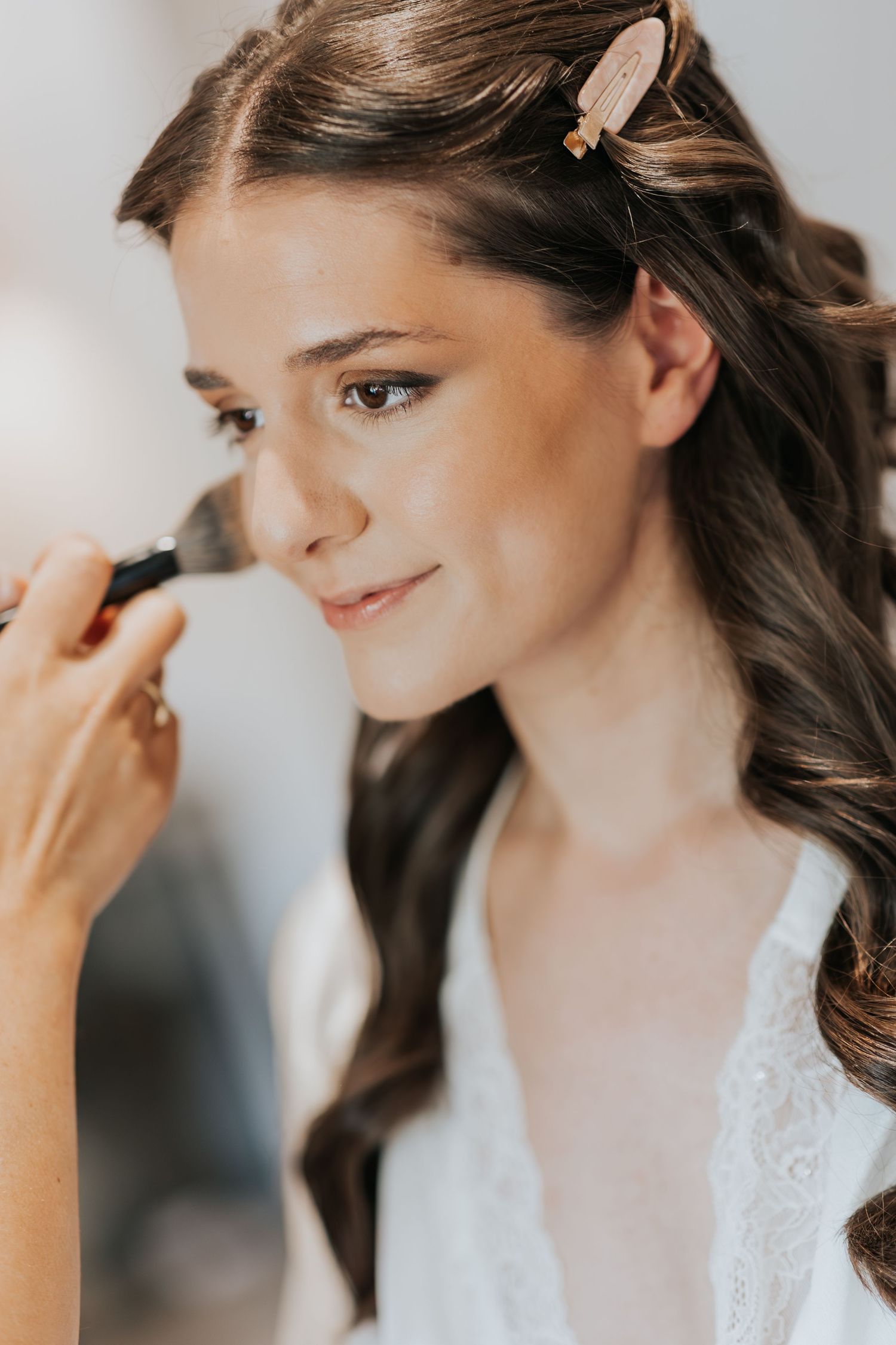 Bride getting makeup applied with a brush, wearing a white lace robe and hair clip, smiling softly.