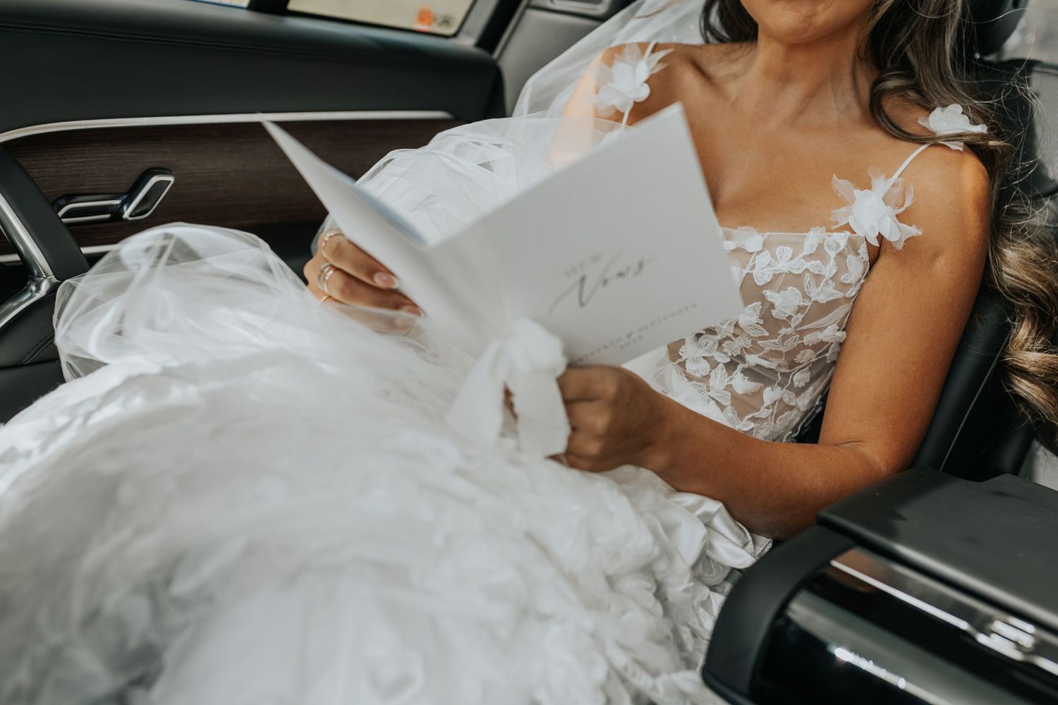 Bride in white lace wedding gown reading a card while seated in a car on her wedding day.