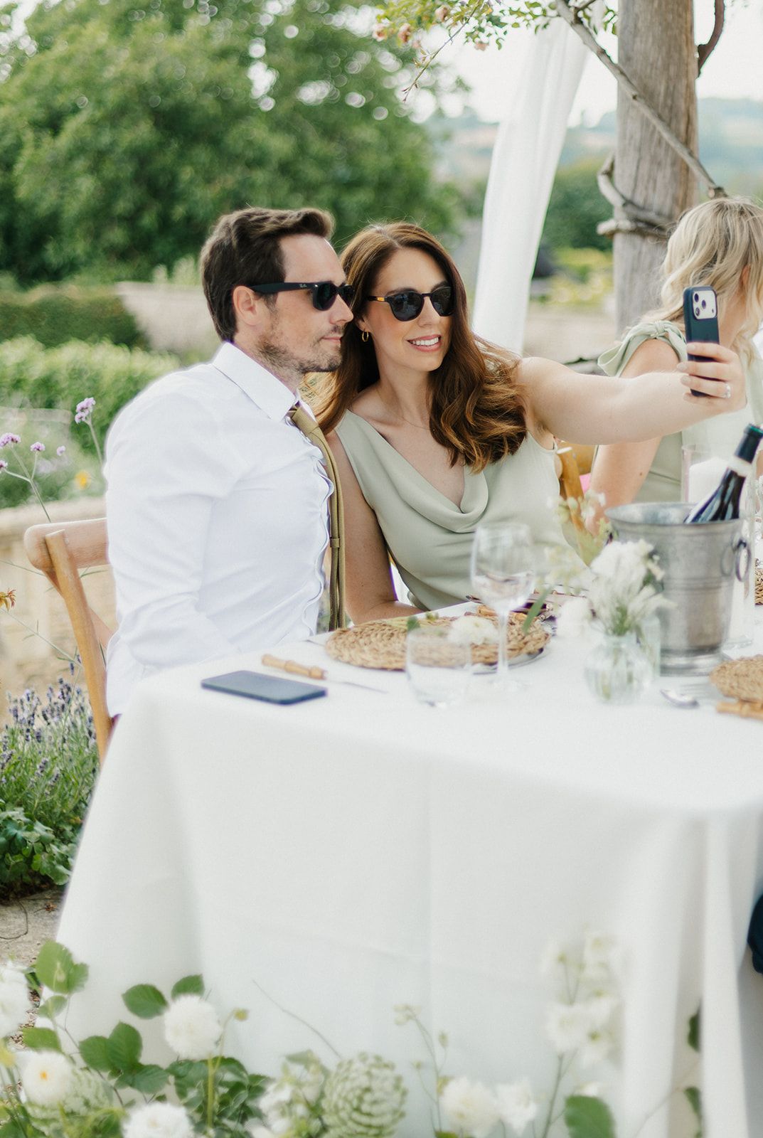 Couple taking a selfie at an outdoor wedding reception table with white florals and greenery.