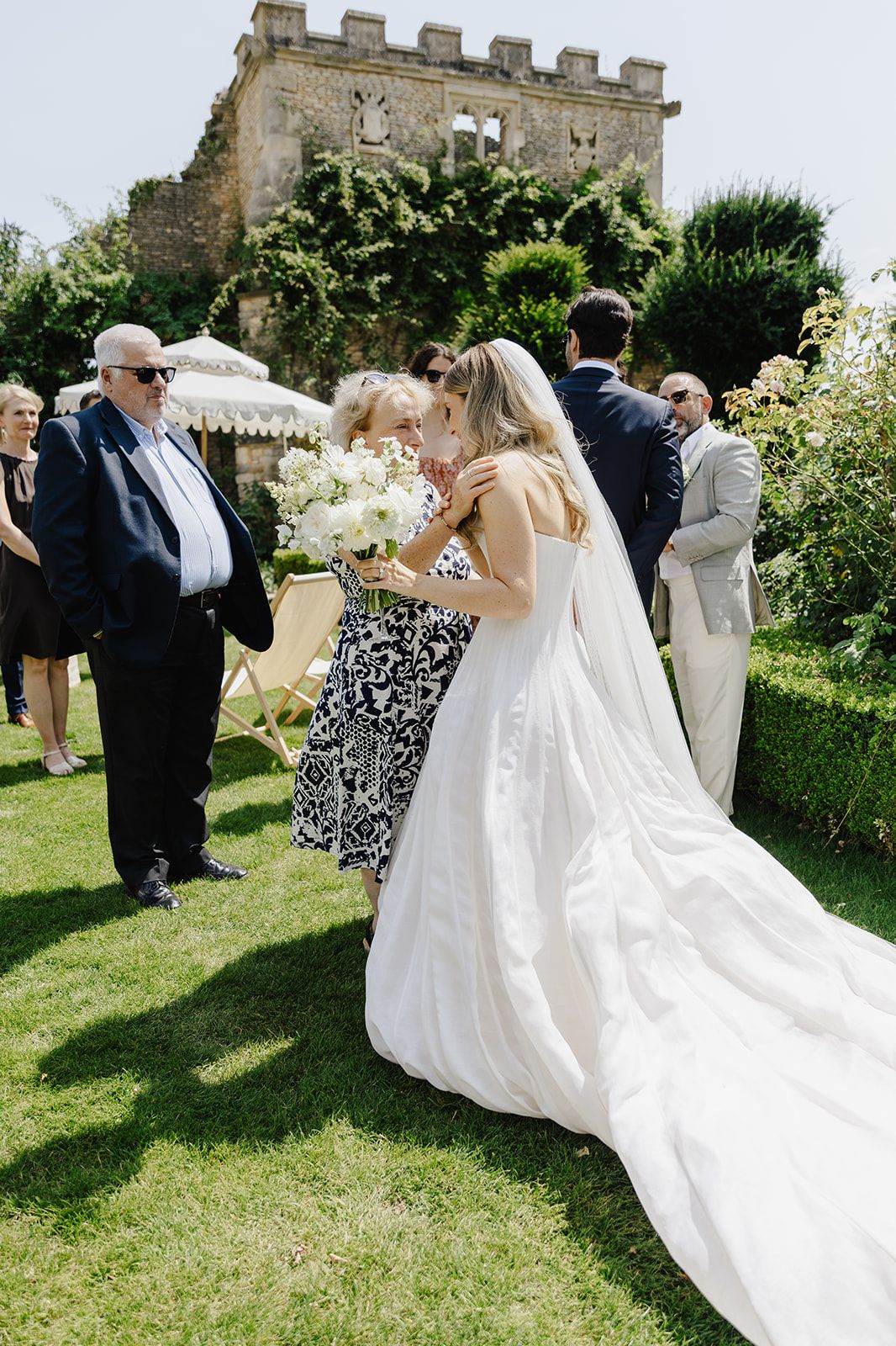 Bride in white gown embraces guest while holding bouquet at outdoor garden wedding near stone castle.