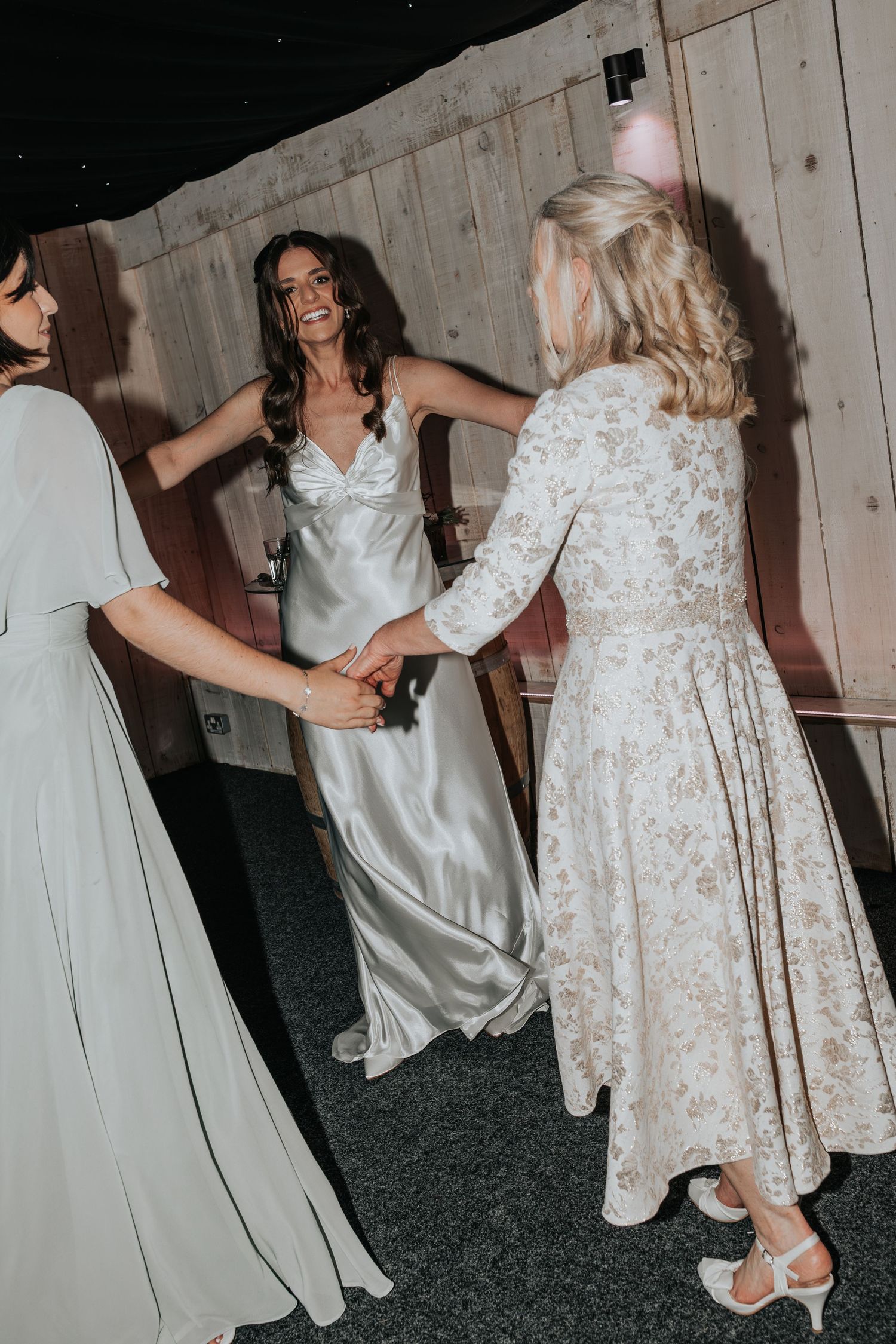 Bride in silver gown dancing joyfully with guests in elegant dresses at a rustic wedding reception.
