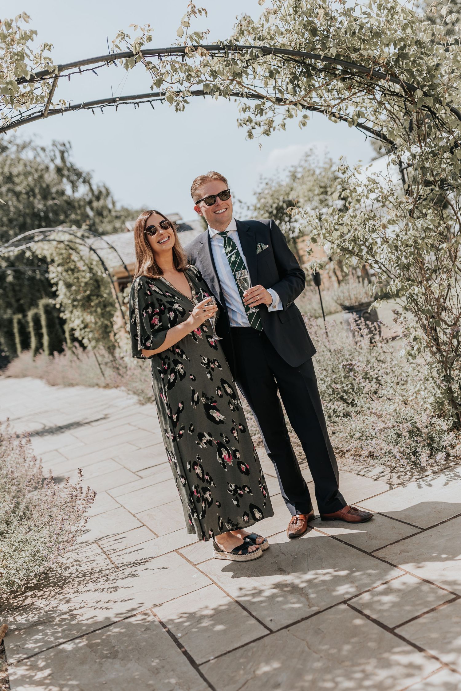 Couple in formal attire smiling under a garden arch, holding champagne glasses on a sunny patio.