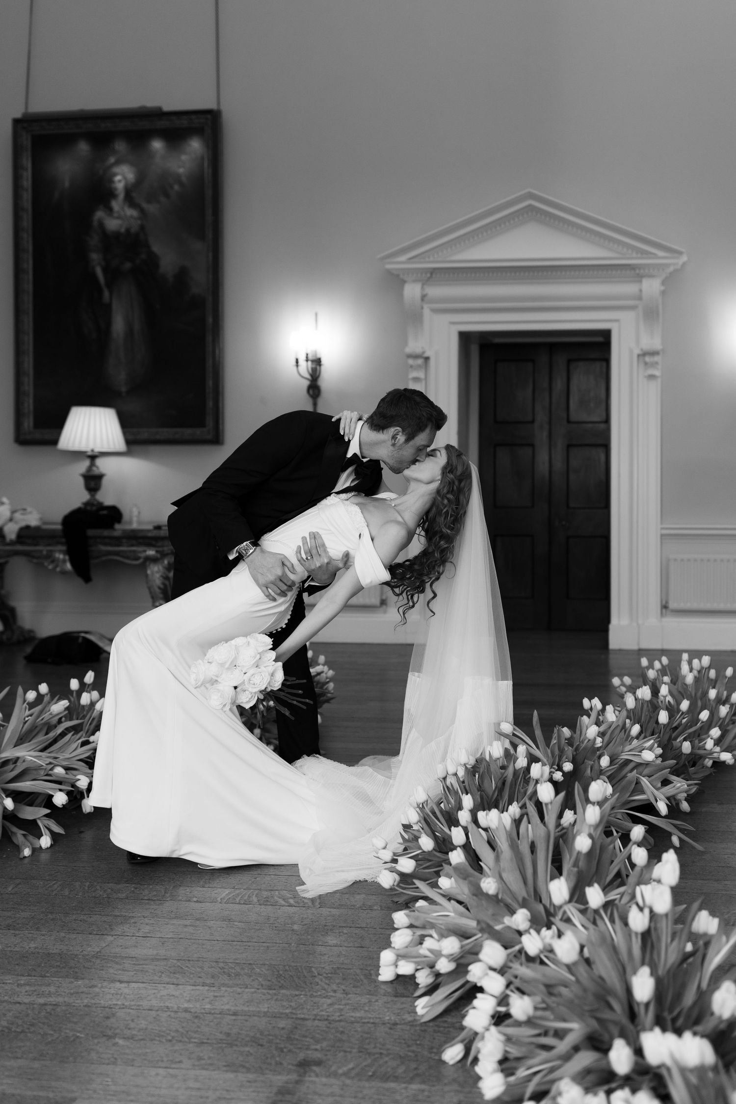 A black and white photograph of a bride and groom embracing in a formal indoor setting with flowers and decorations.