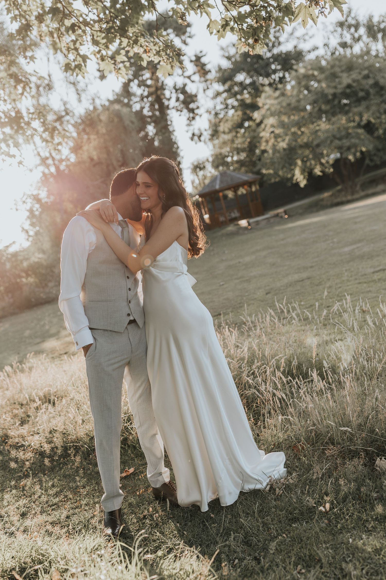 A bride and groom embrace in golden sunset light in a lush green field, wearing elegant white wedding attire.